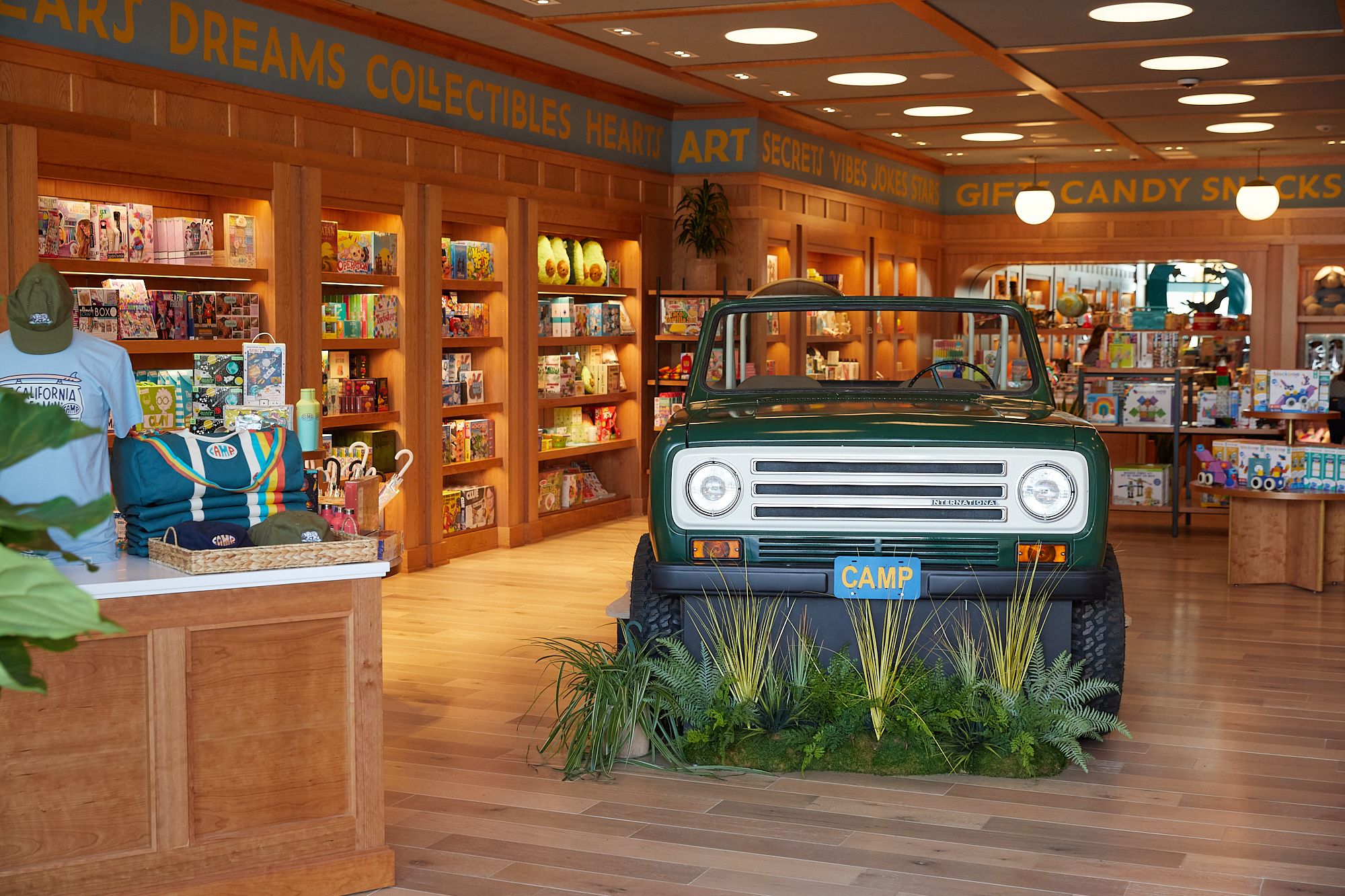 Interior of a brightly lit wooden store with shelves of colorful games and toys, a green vintage vehicle with a "CAMP" plate in the center, plants around the vehicle, and a sales counter with merch on the left.