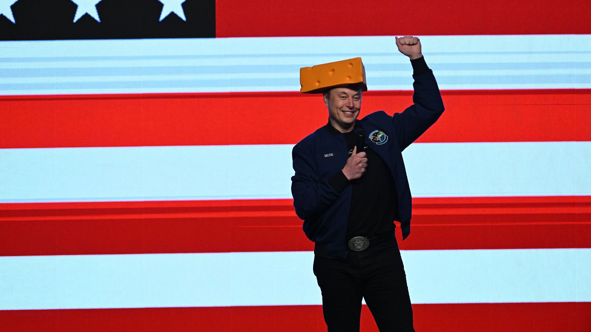 Elon Musk wearing a cheese hat and holding up his fist while standing in front of an American flag backdrop.