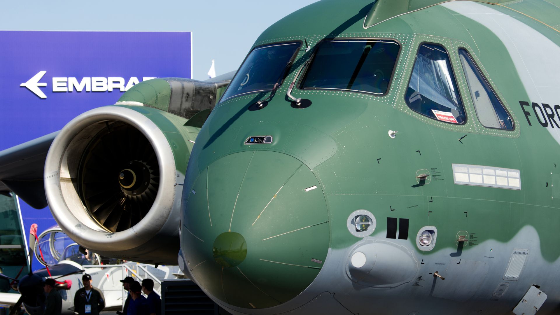 A large green-and-grey military transport aircraft sits in front of a building labeled "Embraer."