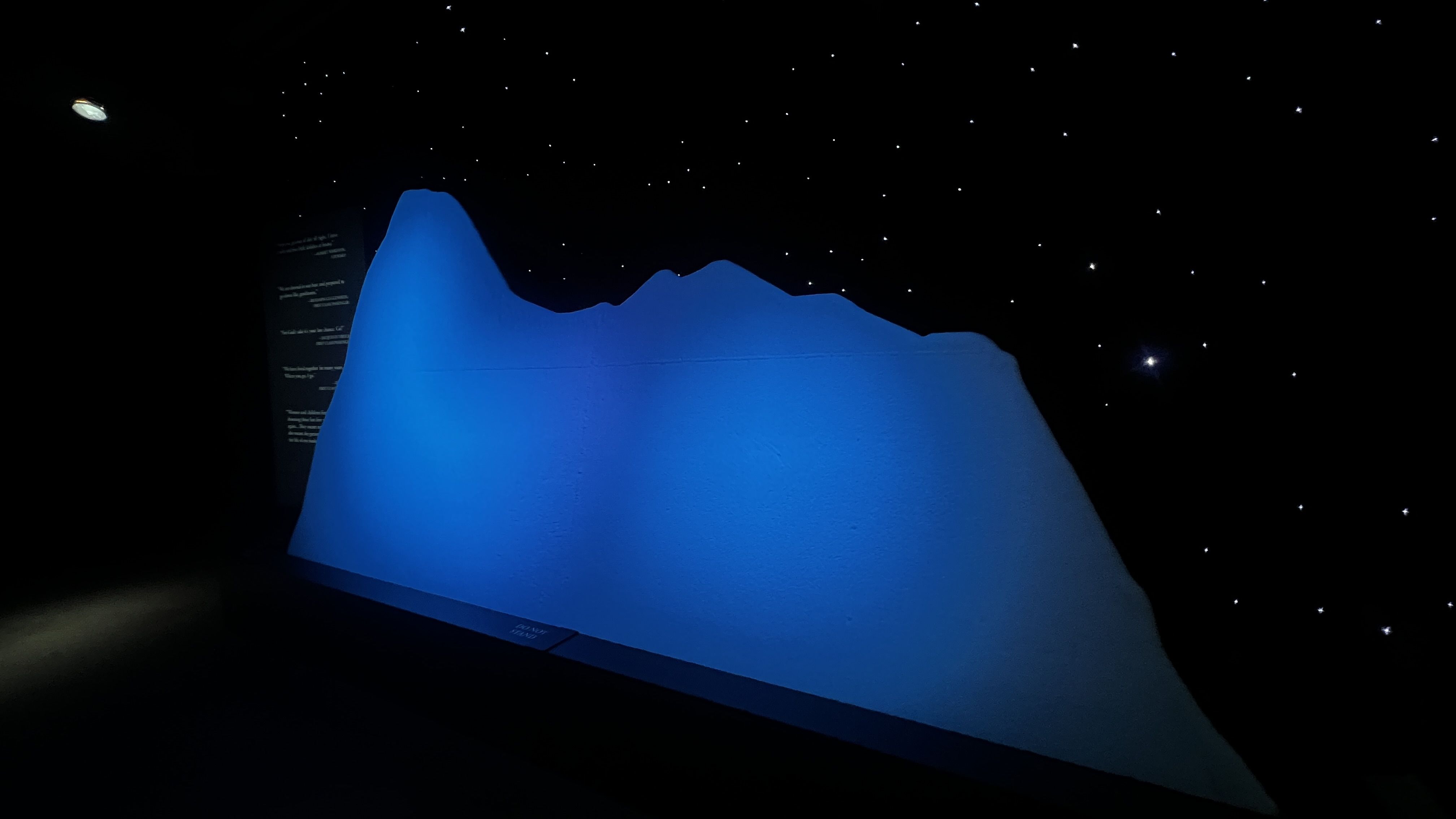 Dim exhibition space with a large blue wavy mound illuminated in blue light; dark starry ceiling with many small white stars; a small round planet-like object visible in the upper left.