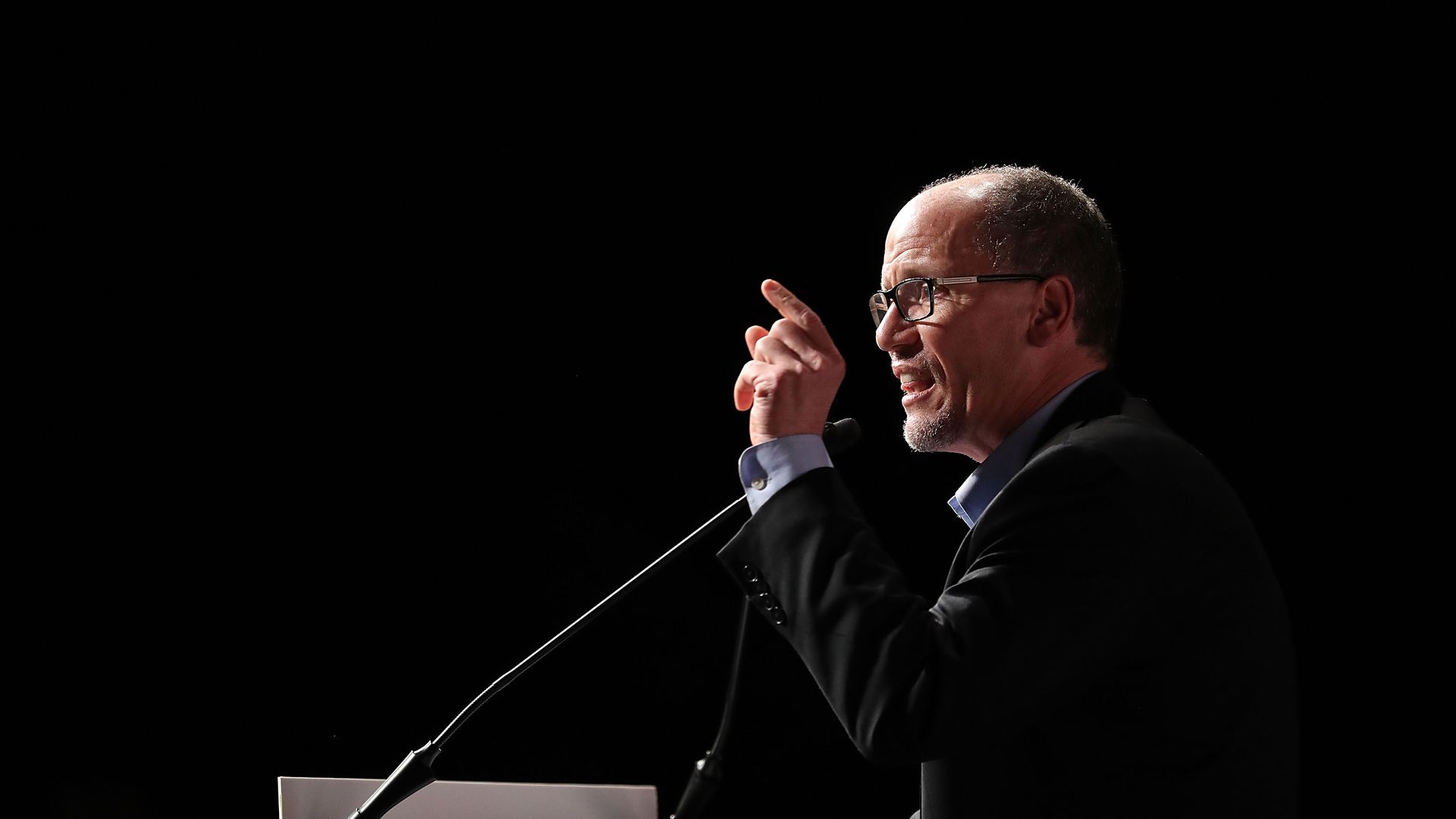 Tom Perez wears black, talks at a podium, and has a light shining on his head, as he points up.