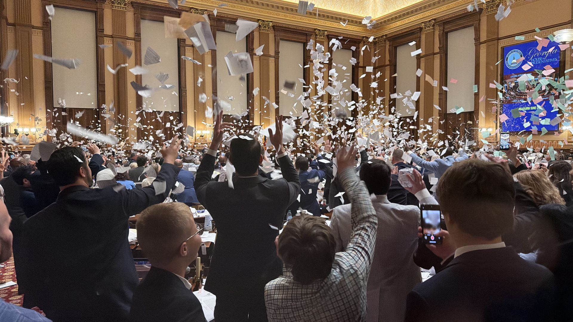 Large, ornate conference hall filled with people in suits celebrating as white confetti rains down; attendees raise arms, some take photos, and a blue banner decorates the far wall.