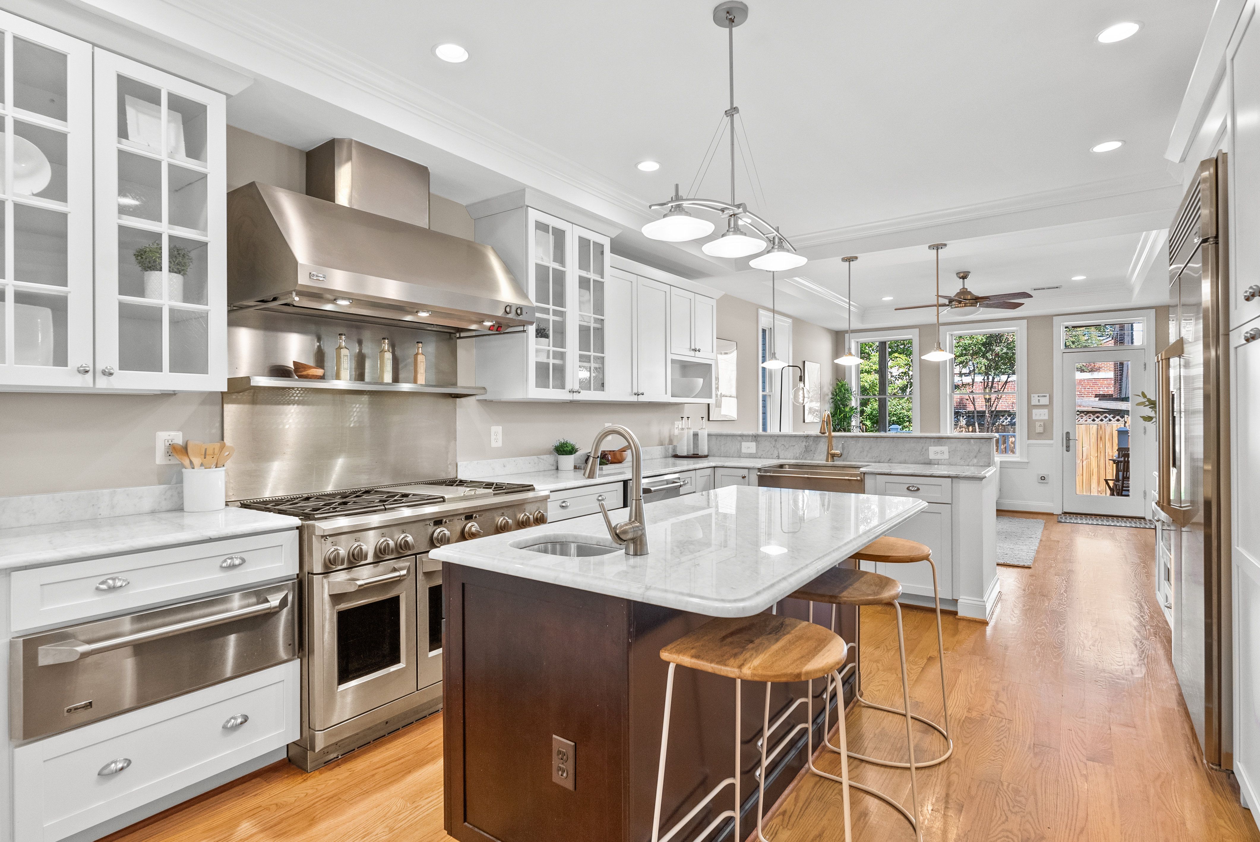 Modern kitchen with white cabinets, stainless steel appliances, wooden floor, pendant lights, and a marble island with two wooden stools, opening to a backyard door and windows.