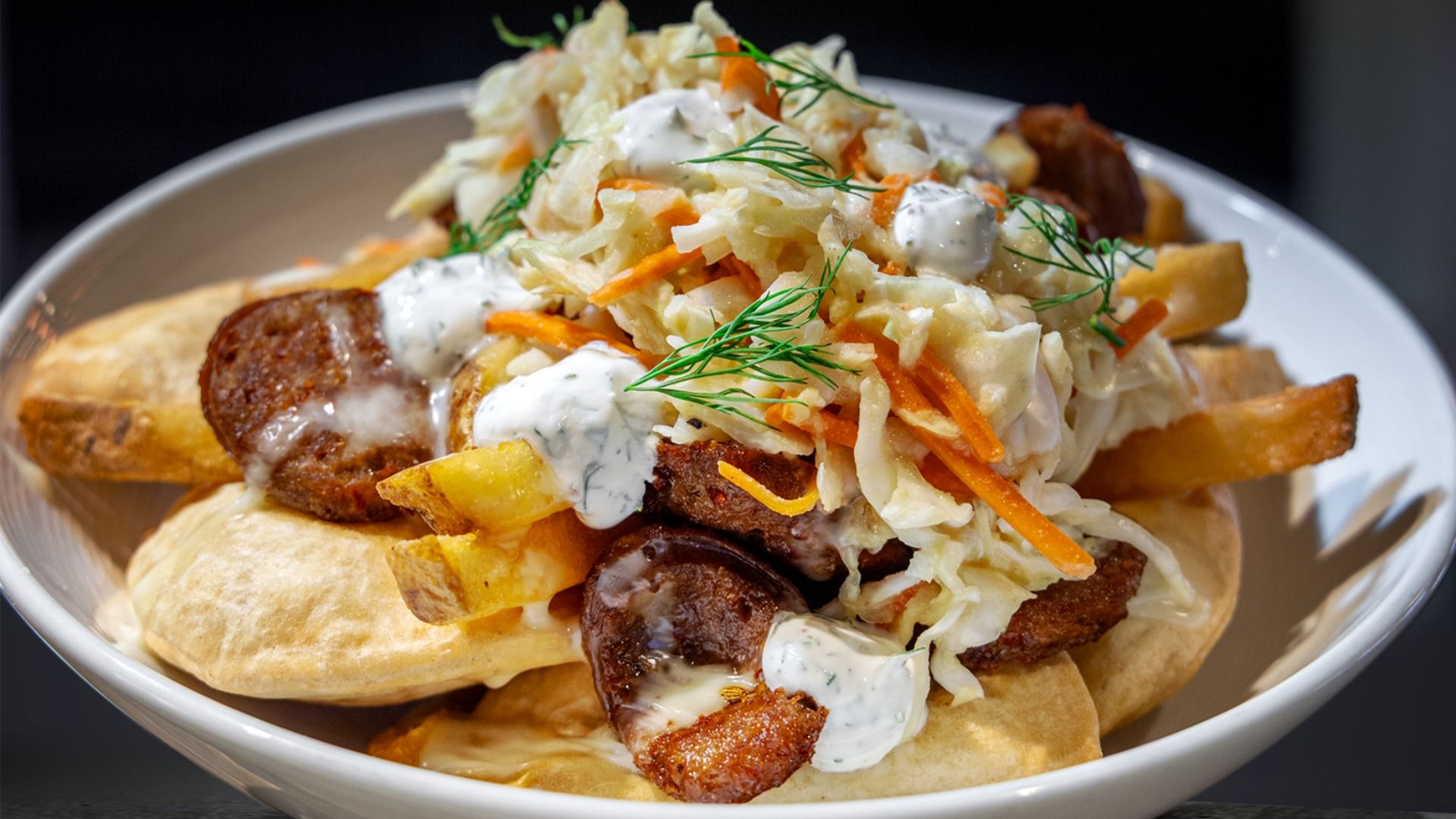 Plate of pirogies topped with fries, Italian sausage slices, shredded cabbage and carrots, creamy white sauce, and fresh dill garnish.