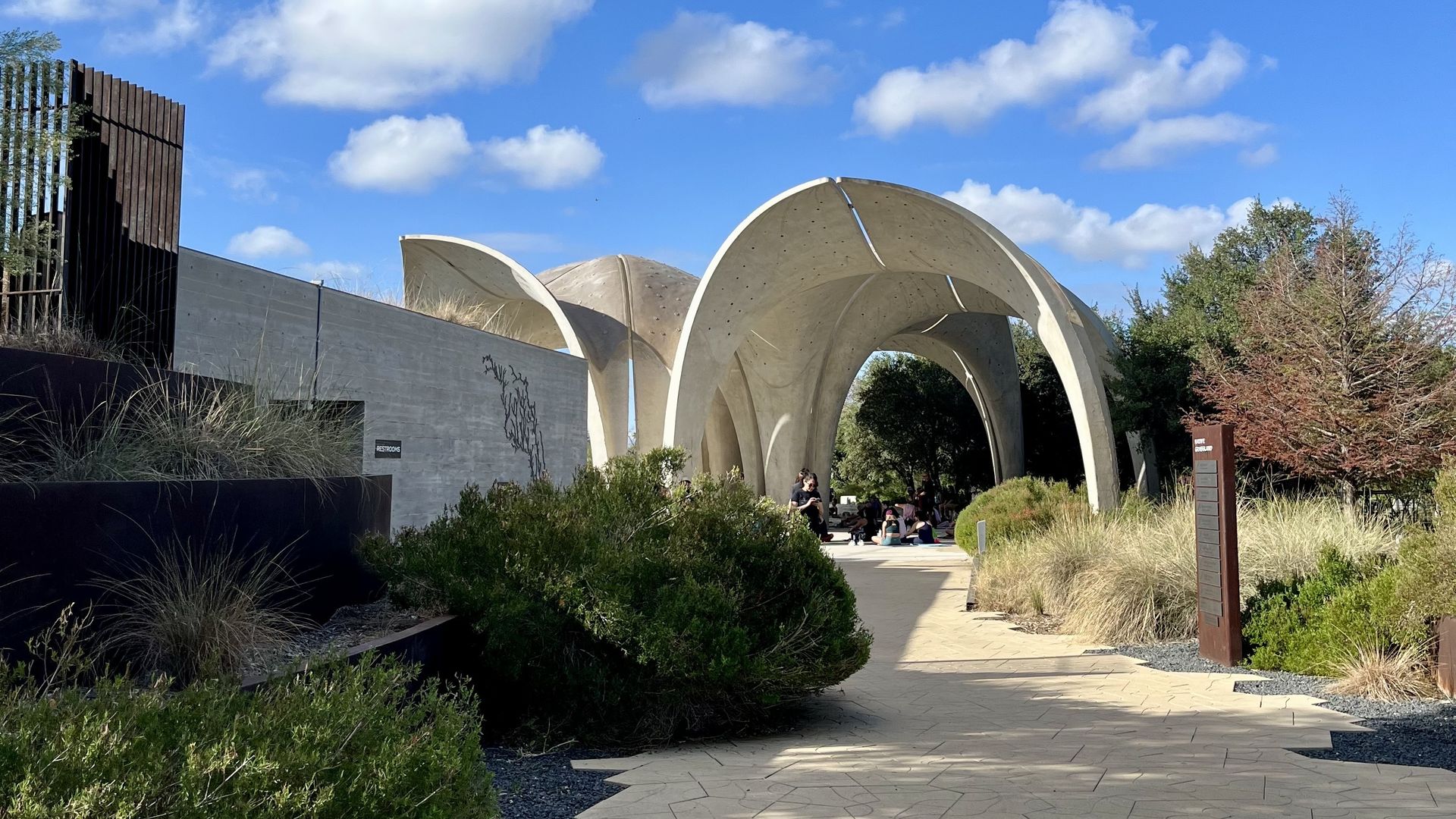 Sunny day at a campus with white curved arches forming a tunnel. People sit beneath. Hexagonal paving, green shrubs along the path, and a blue sky with fluffy clouds.