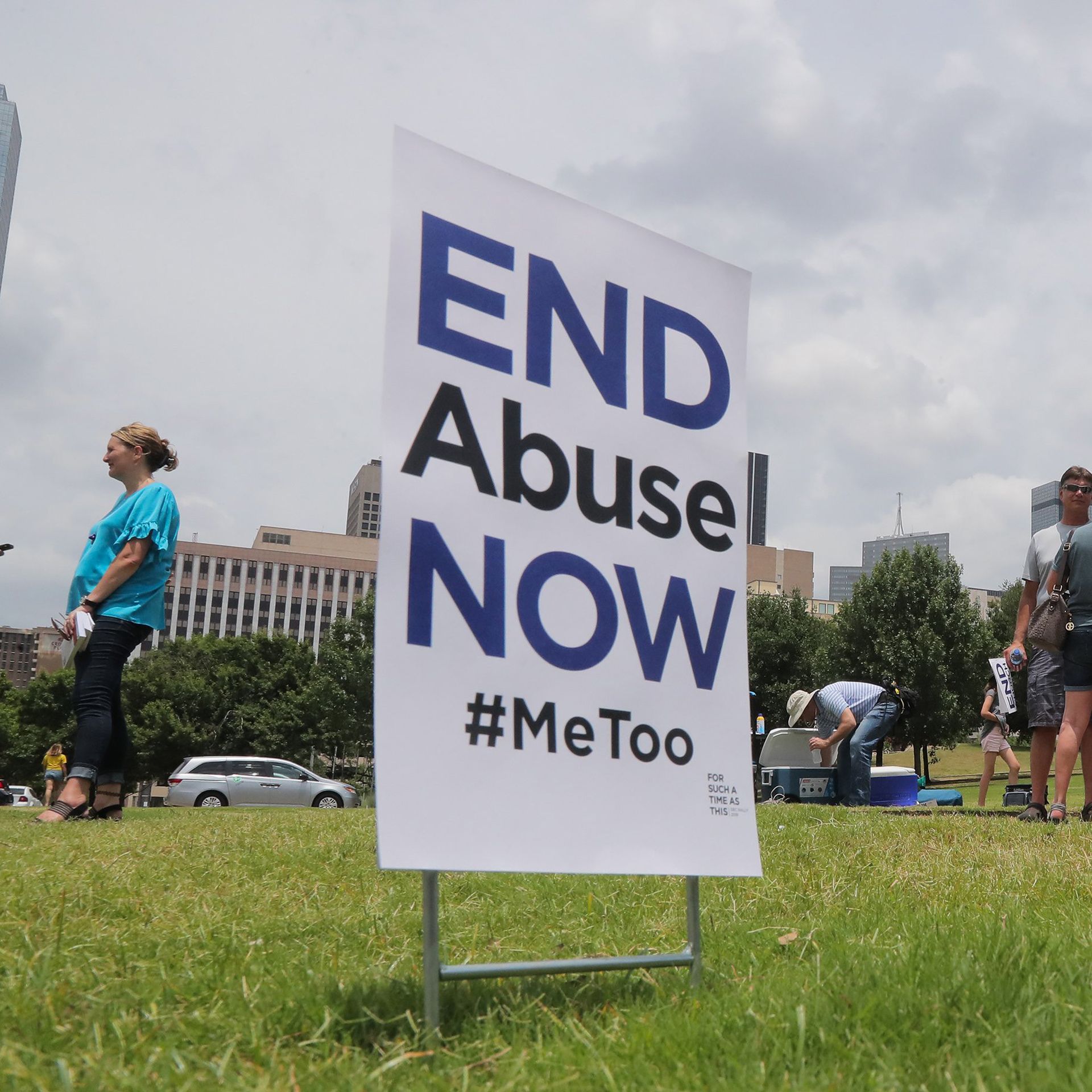 Photo of a sign that says "End Abuse Now #MeToo" embedded in grass with people standing around