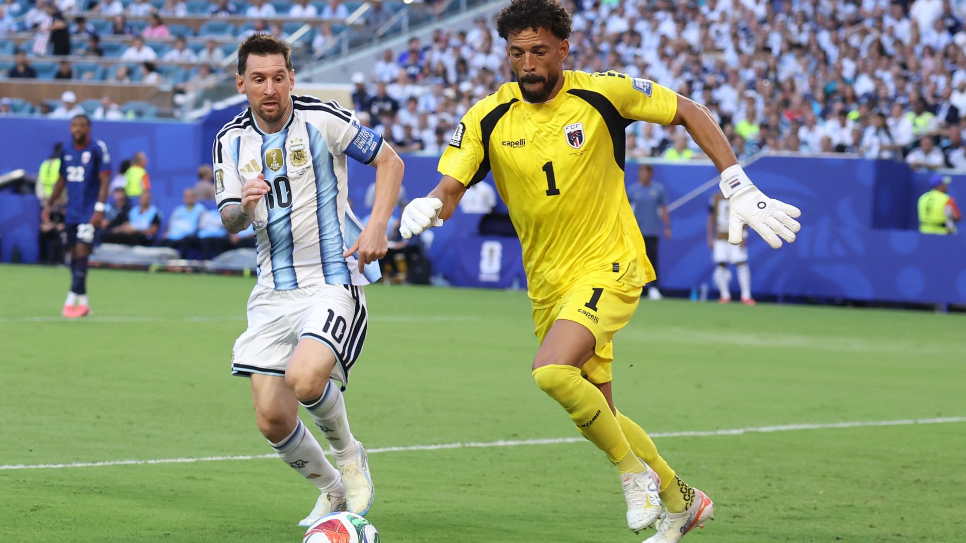Two soccer players chase after a ball on the field. One player wears white shirts, and a blue and white striped shirt. The other player wears yellow shorts and short with the number 1 on the front. 