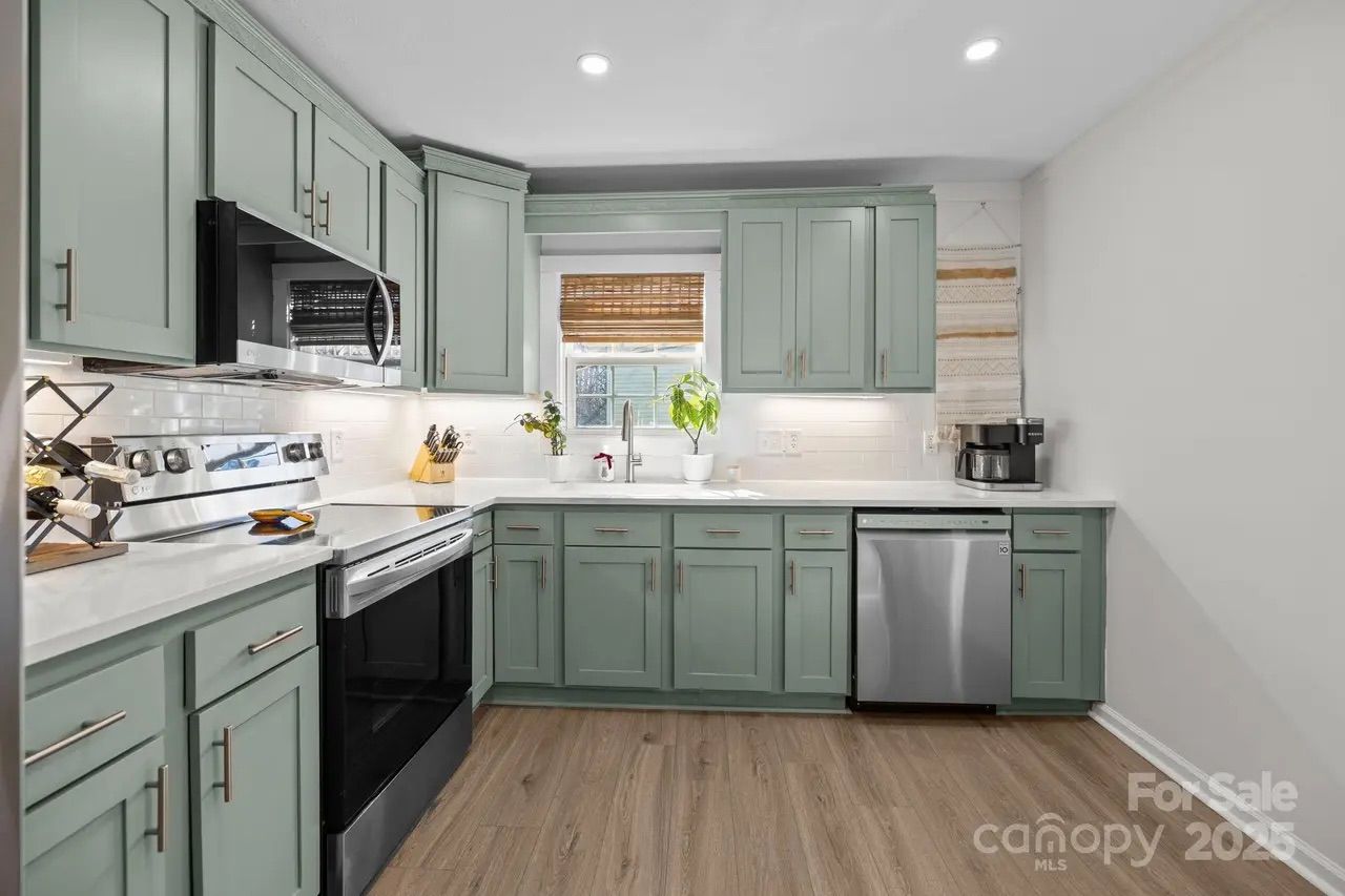 Modern kitchen with sage green cabinets, white countertops, stainless steel oven, microwave, and dishwasher, wooden floor, window with bamboo blind, potted plants, and coffee maker on counter.