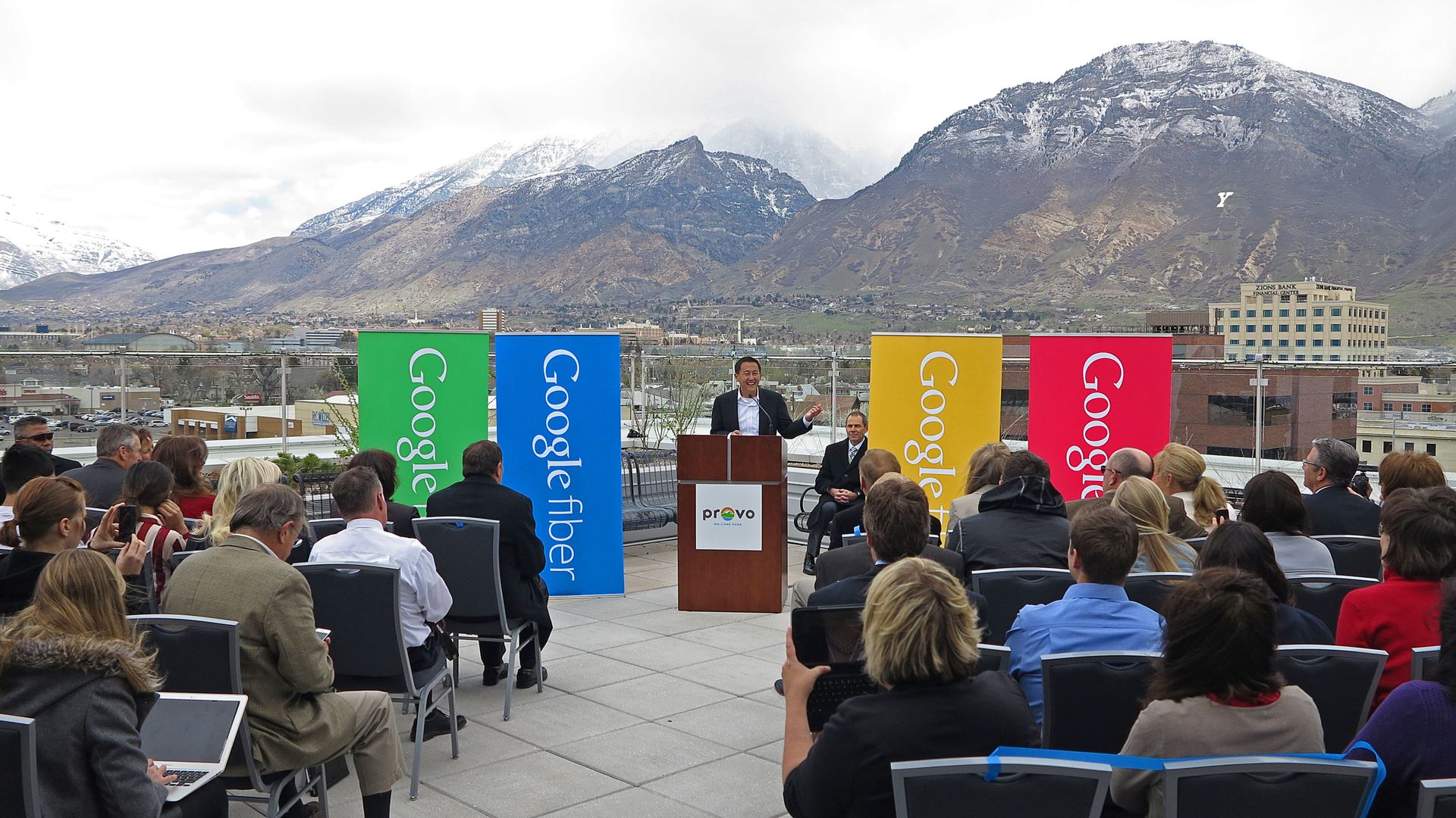 A man speaks in front of banners that say "Google" on a rooftop overlooking Provo, Utah with mountains in the background.