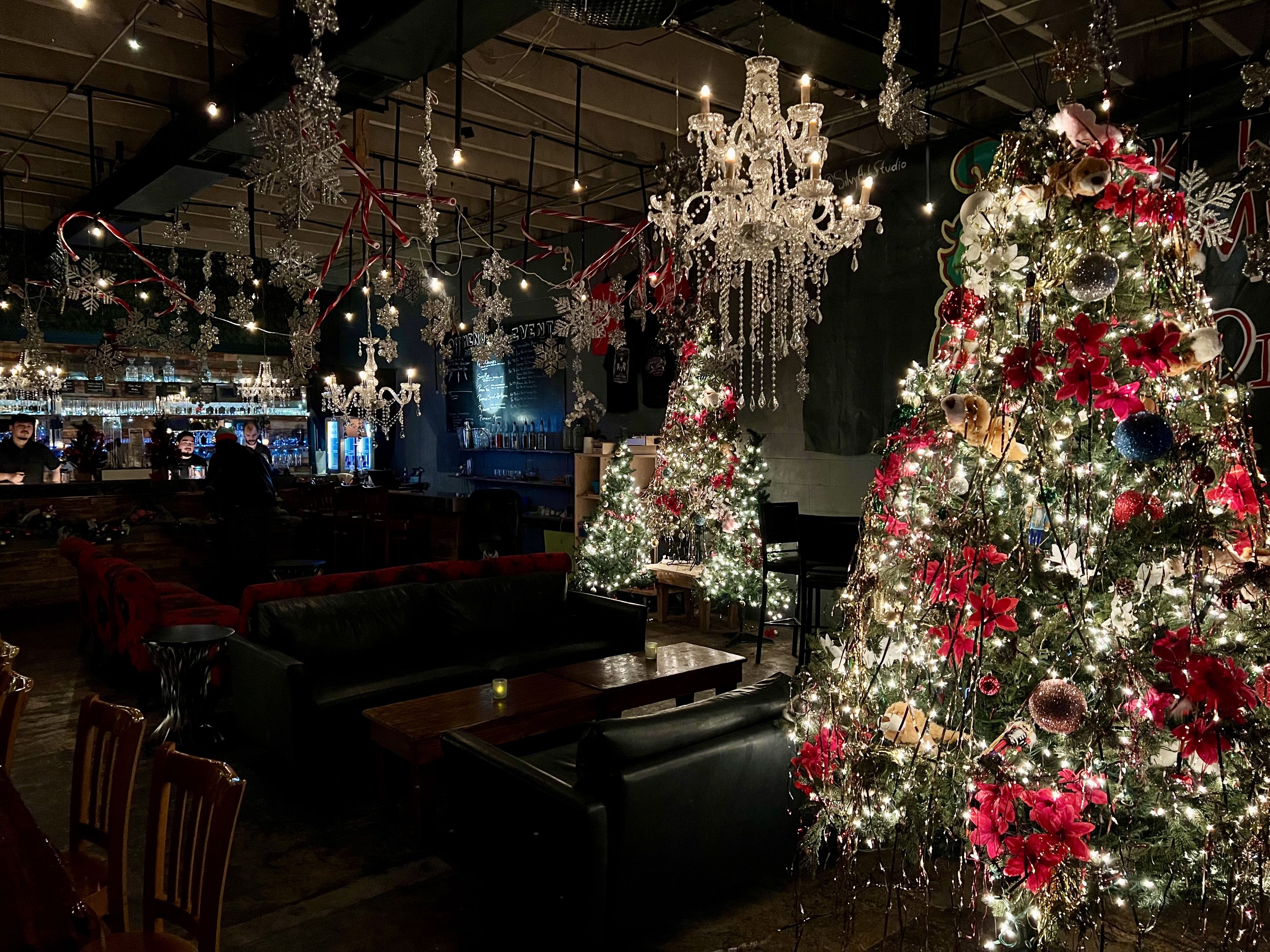 Dimly lit bar with multiple decorated Christmas trees featuring red flowers and lights, chandeliers with candles, hanging candy canes and snowflakes, and cozy black leather sofas.