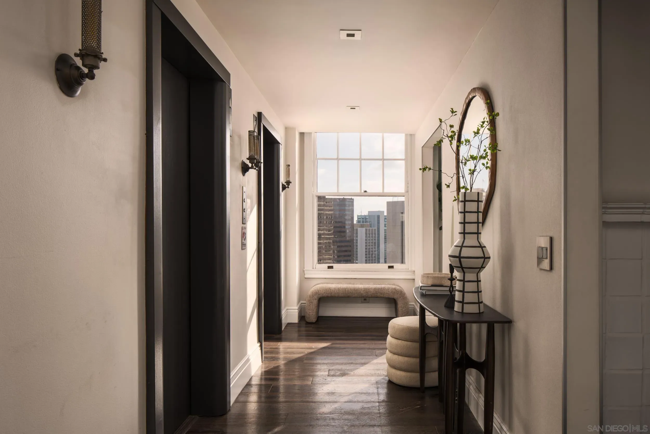 Hallway in a city apartment with dark doors on the left, vintage wall sconces, and a large window at the end showing the skyline. On the right, a tall striped vase with greenery beside a round mirror.