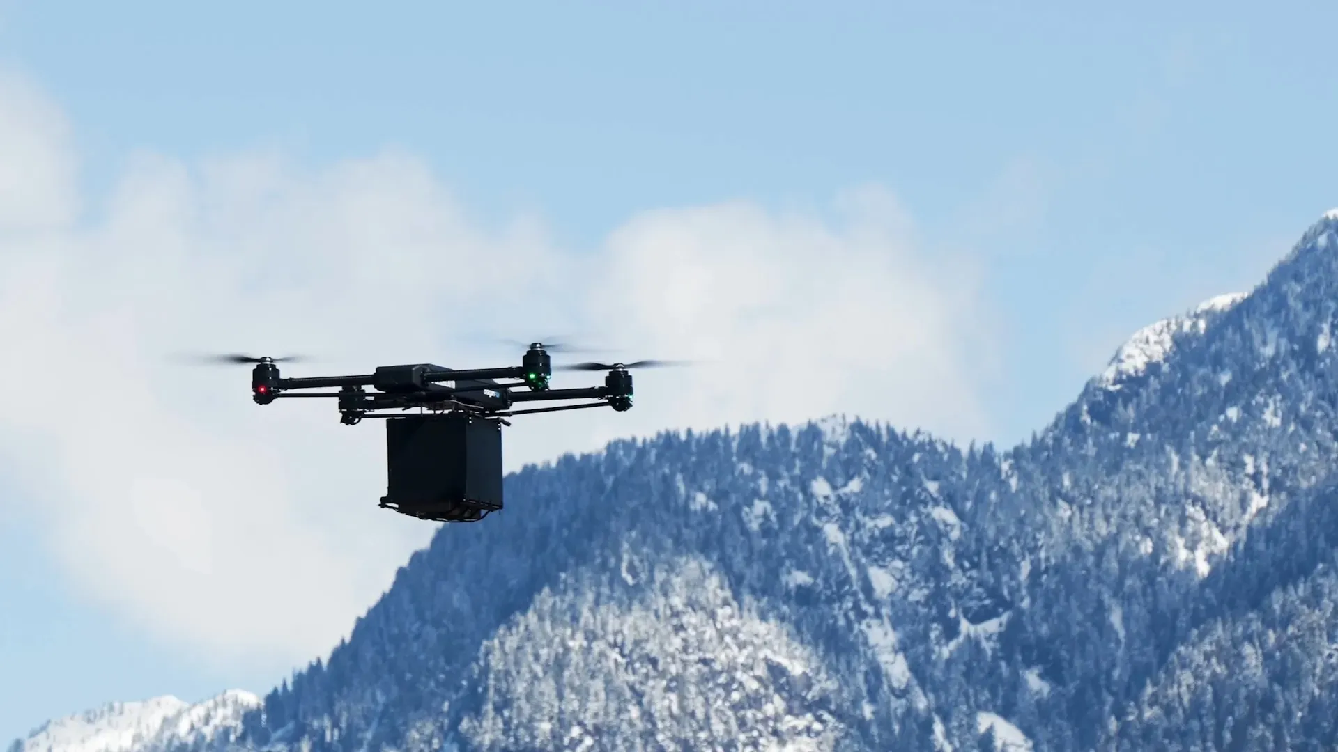 Black delivery drone flying with a box under it against a backdrop of snow-covered mountains and blue sky with clouds.
