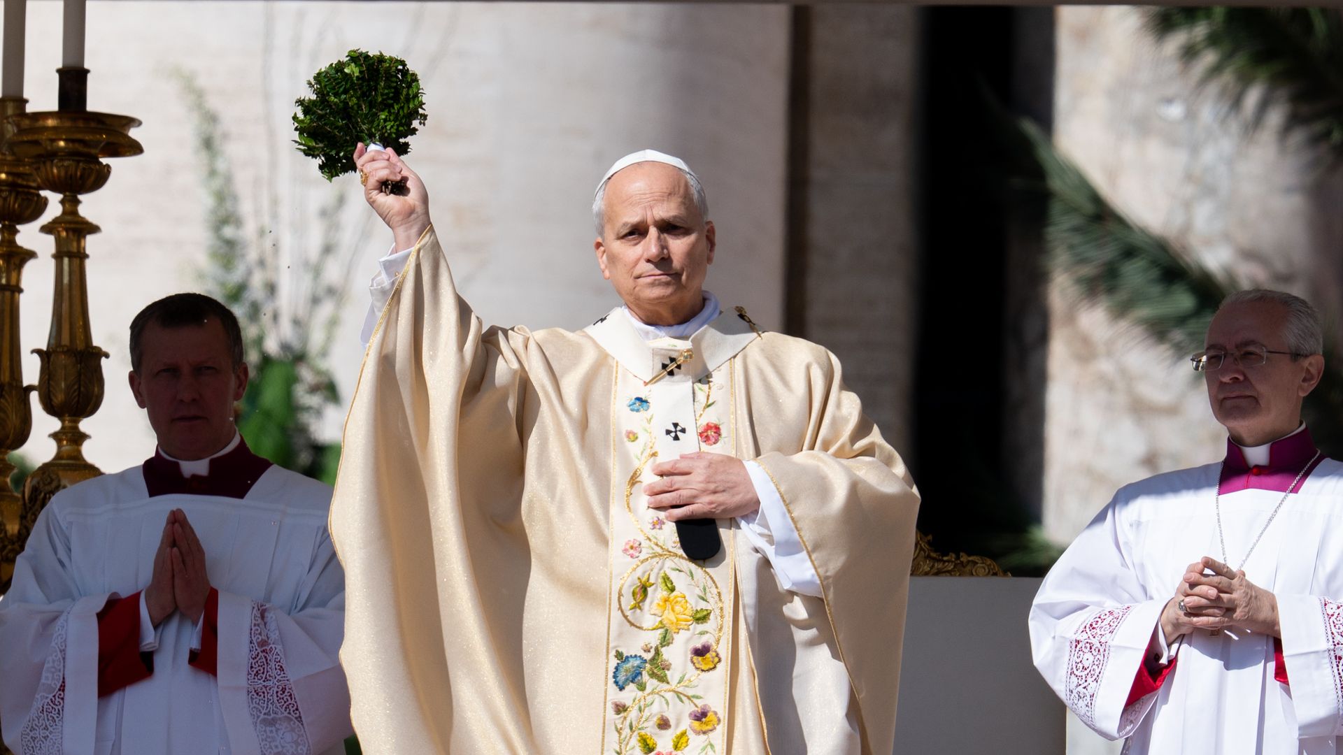 Pope Leo XIV, an older man in cream vestments with a floral-embroidered stole raises a small green wreath, flanked by two priests in white robes during an outdoor religious ceremony.