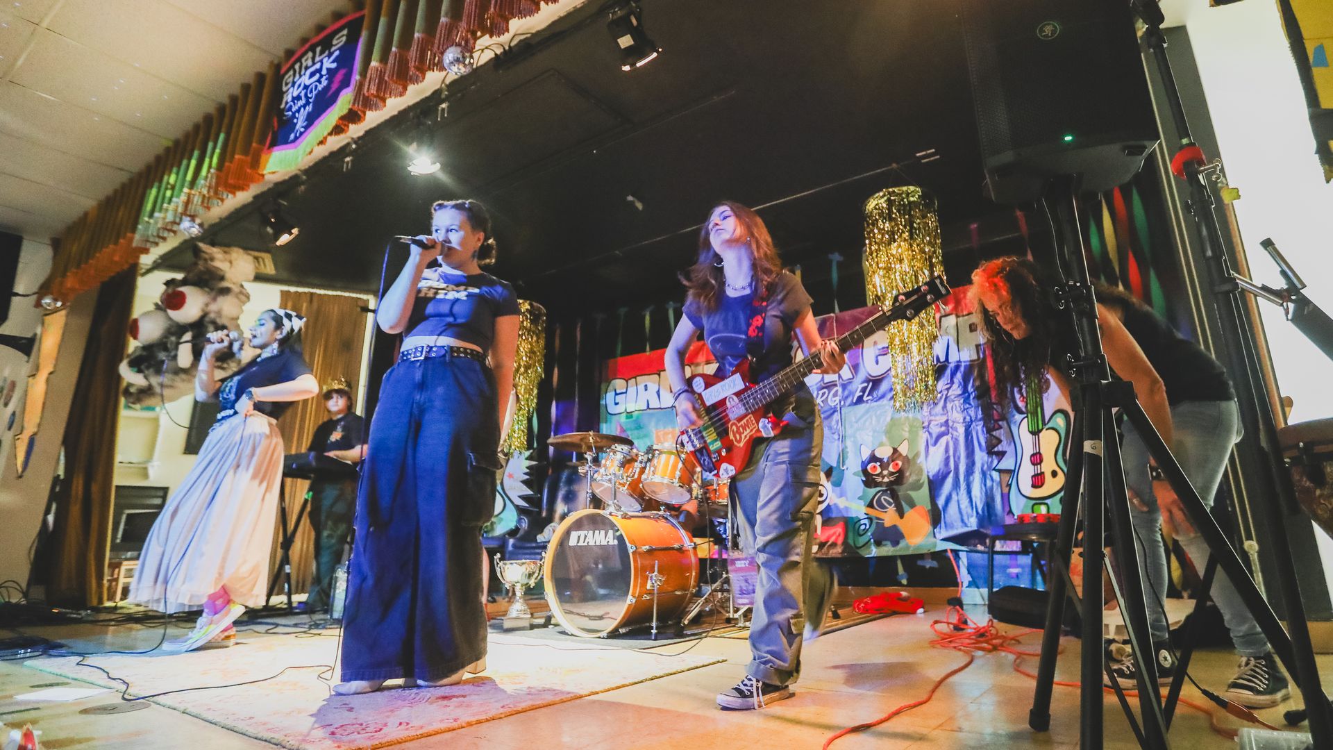  band of teens performs under a "girls rock st pete" banner. The lede singer is wearing shirt that says "dump him"