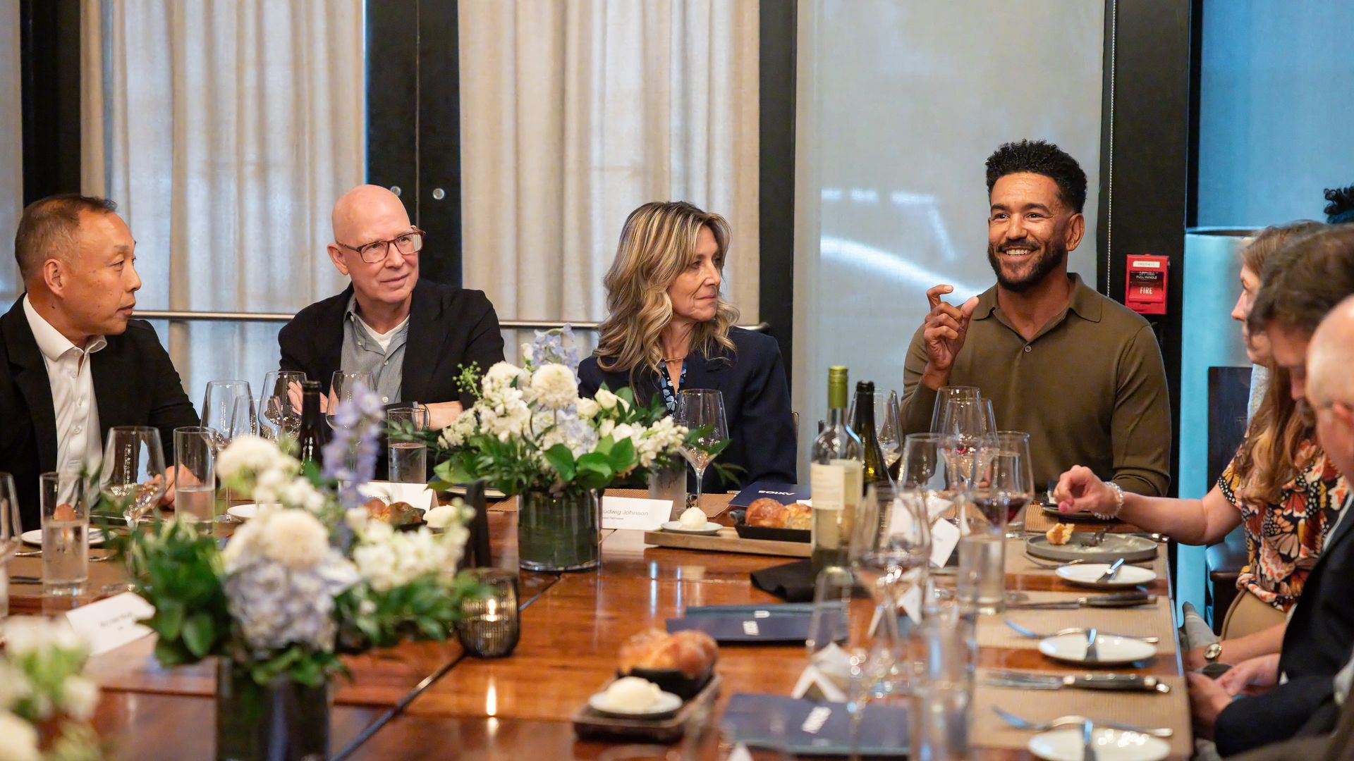 Adults sit at a long banquet table at a formal event, chatting and smiling. Floral centerpieces, wine glasses, and bread on the table with beige curtains and a blue wall behind.