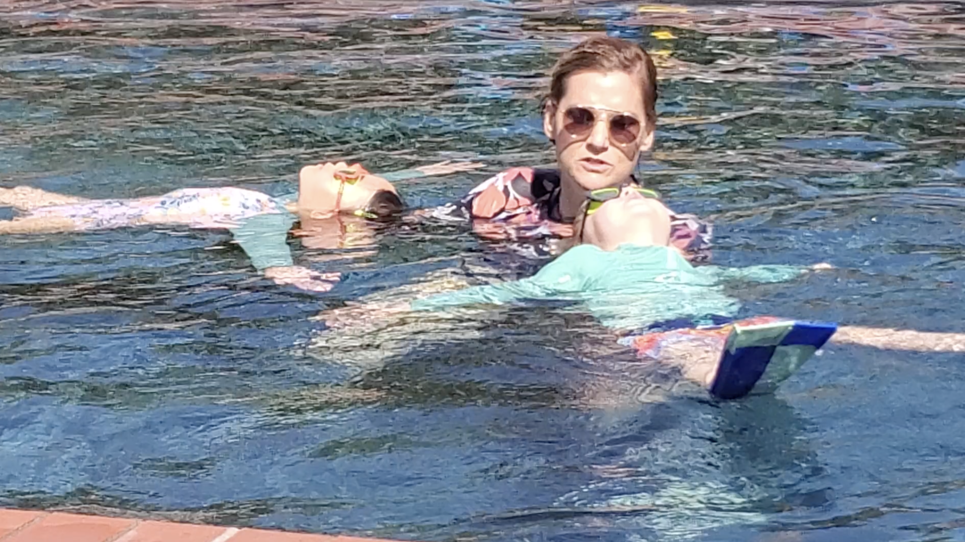 Two people swimming in clear blue water by the pool: a woman wearing aviator sunglasses and a pink patterned shirt holds a child in turquoise, both floating near the surface.