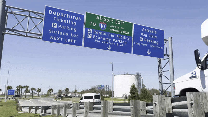 Overhead blue airport signs direct departures, arrivals, and the airport exit. A white van and a white truck pass below on a clear day, with palm trees, a wooden guardrail, and a blue sky.