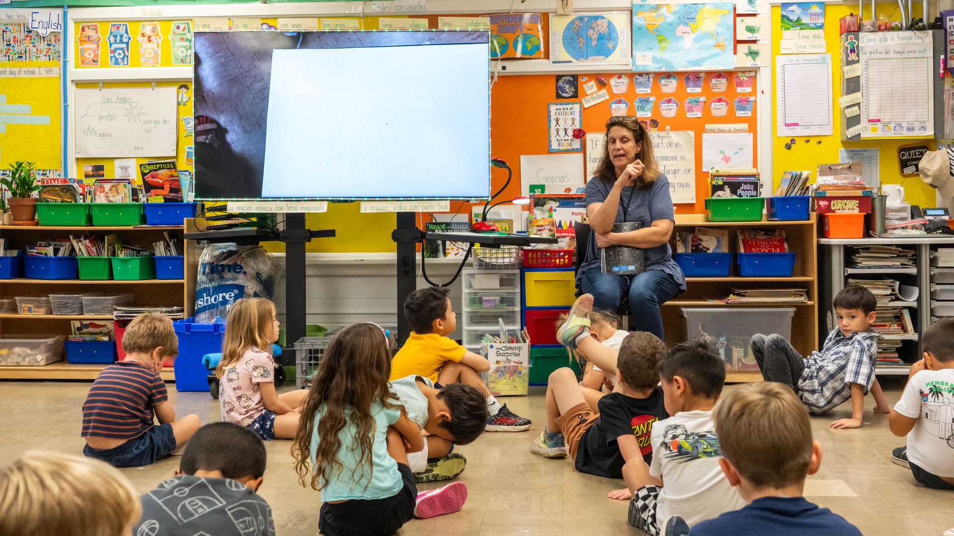 A teacher sits in a chair with little kids on the ground in front of her in an elementary school classroom.