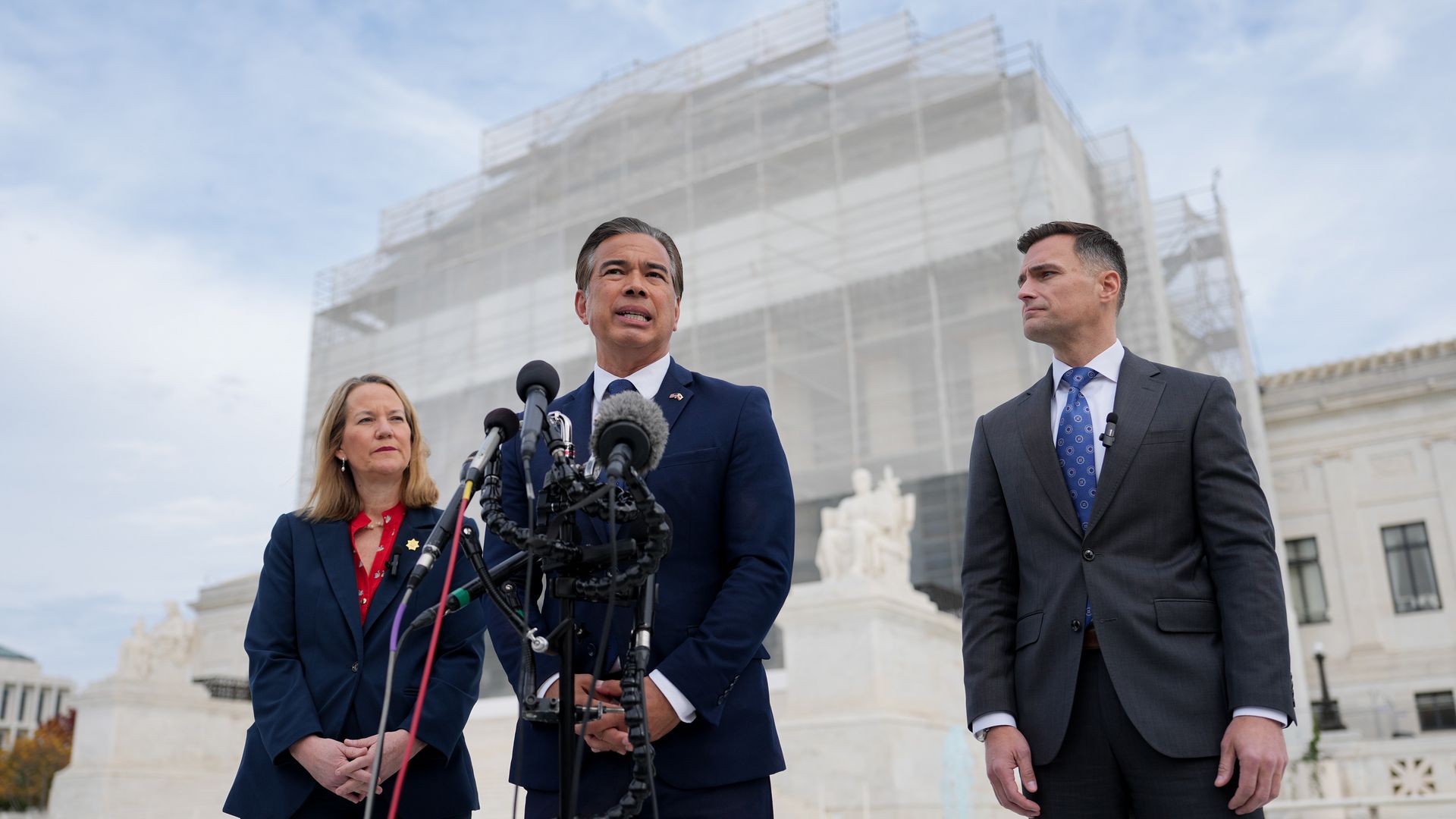 A group of people in suits in front of a group of microphones, with a building behind them wrapped in scaffolding and mesh.
