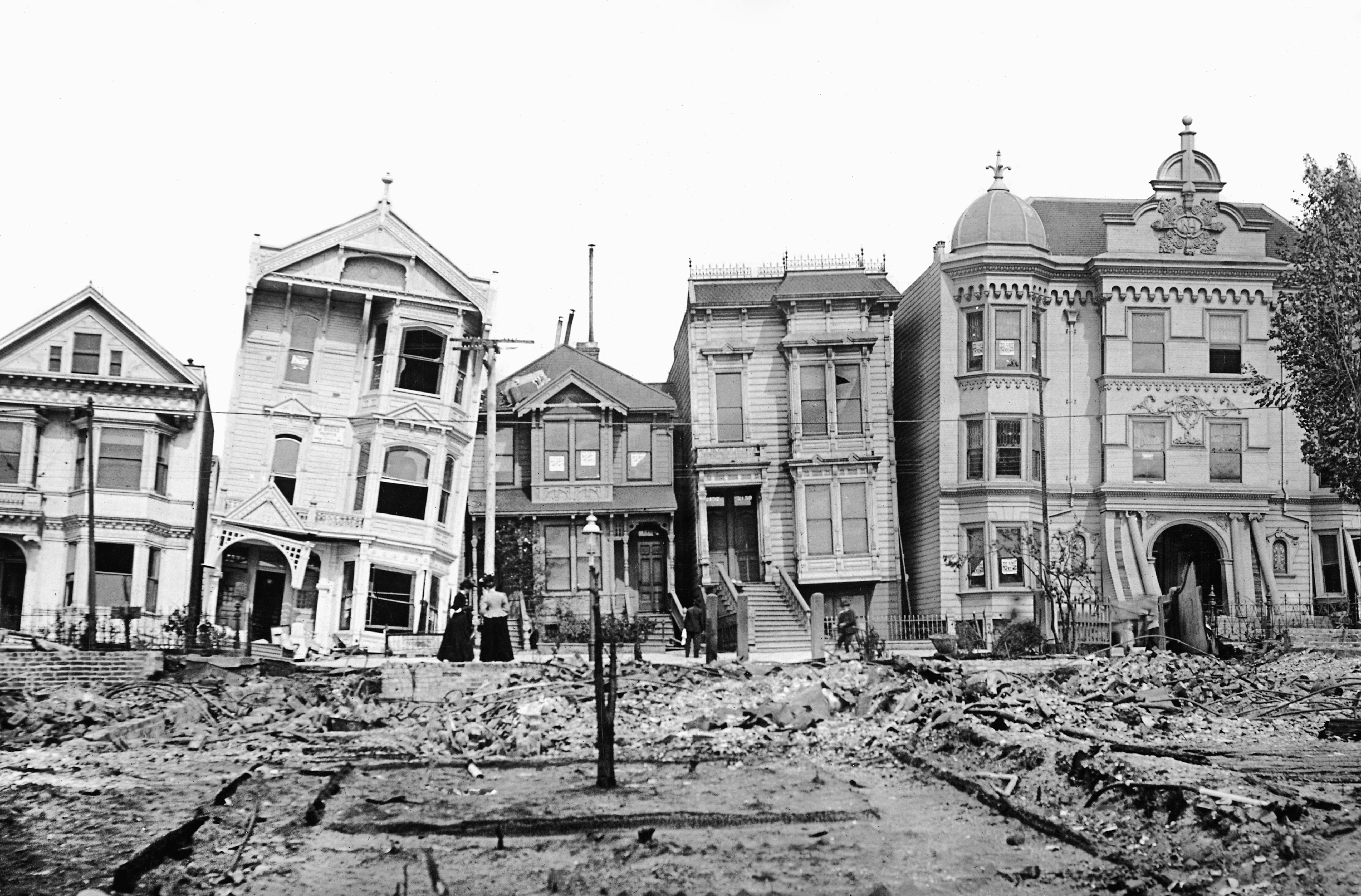Black-and-white photo of a row of ornate Victorian houses, some tilted or damaged, with rubble in the foreground and a few people standing along the street.
