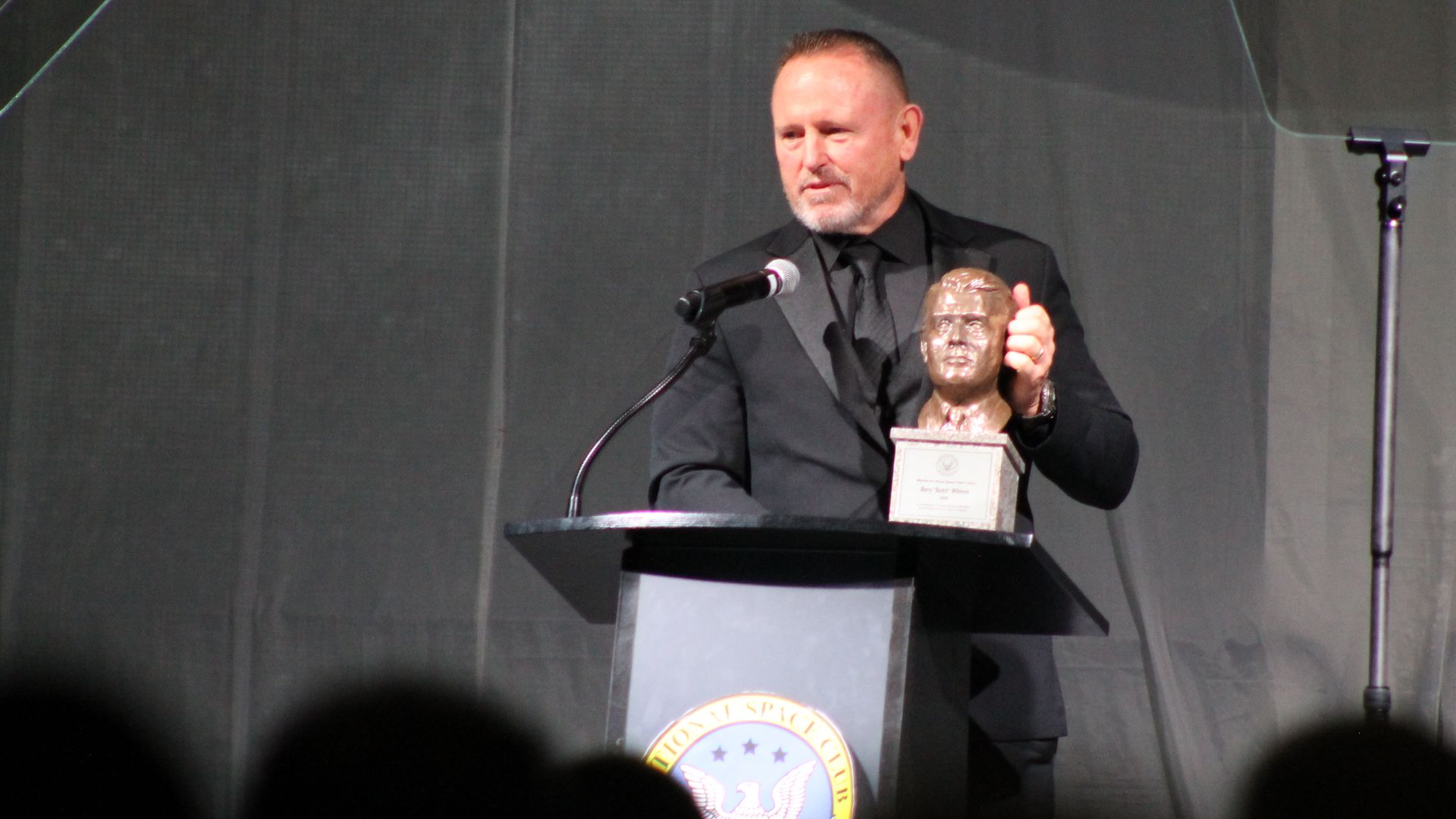 Man in black suit stands at podium with National Space Club emblem, holding a bust trophy, speaking into microphone with teleprompters on each side against dark backdrop.