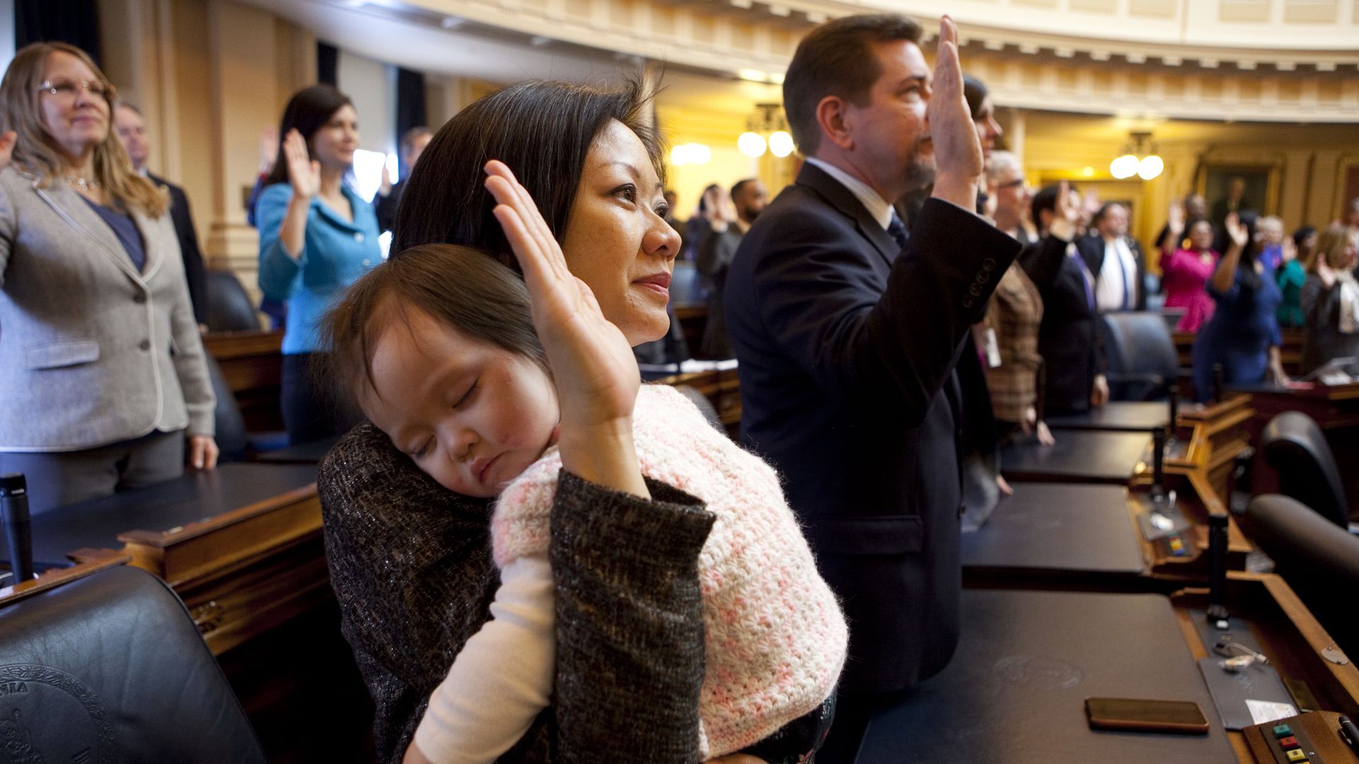 A woman holding a sleeping baby while her right hand is up and about to be sworn in at the state legislature next to other politicians also being sworn in with their right hand up