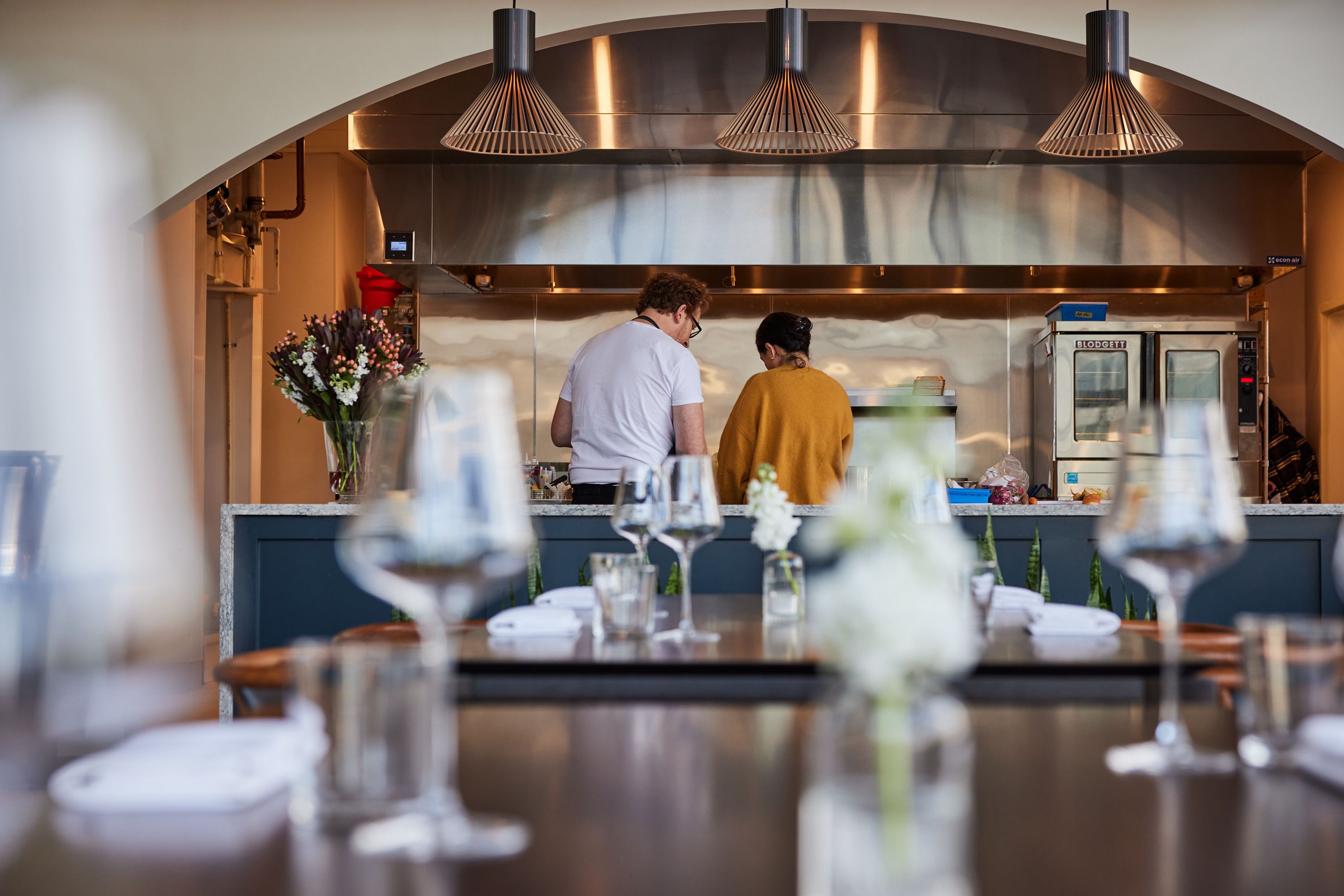 Two people, a man in a white shirt and a woman in a mustard sweater, work at a stainless steel counter under three cone pendant lamps in a kitchen; blurred tables with flowers in foreground.
