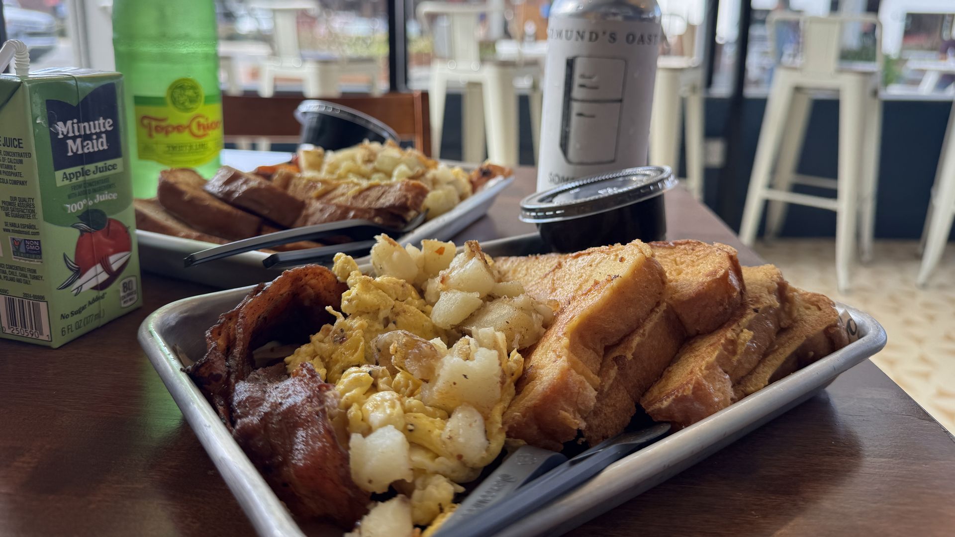 Two breakfast trays on a wooden table with bacon, scrambled eggs, potatoes, and toast; a green Topo Chico bottle, a Minute Maid apple juice box, and a silver can in a bright cafe with white stools.