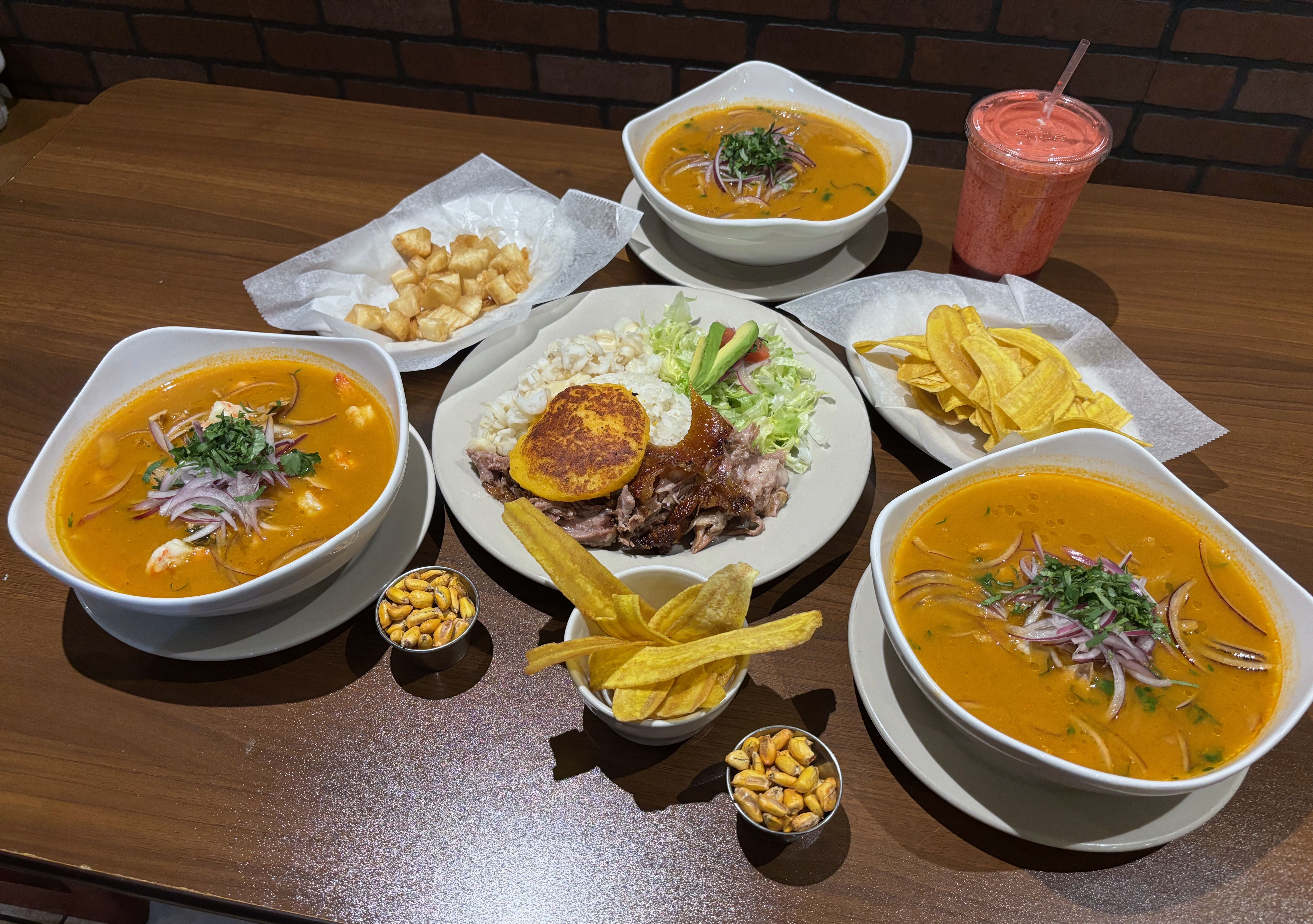 An overview of three bowls of encebollado and hornado in the center, with fried yuca and plantain chips and cancha cups surrounding it