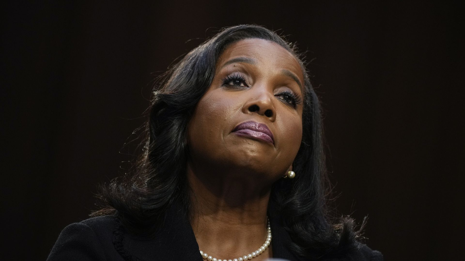 Close-up of a woman with dark hair, pearl earrings, and necklace, wearing a black top, looking upward with a serious expression against a dark background.