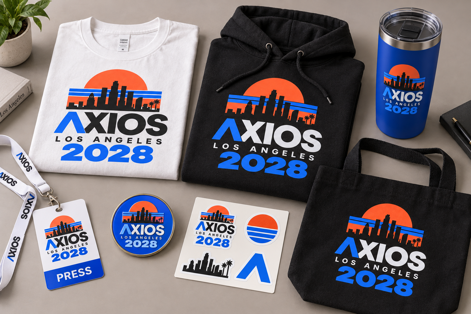 Axios Los Angeles 2028 merchandise: white and black tees with an orange sunset skyline and blue "AXIOS 2028" text, plus a blue tumbler, tote, lanyards, badges, and stickers.
