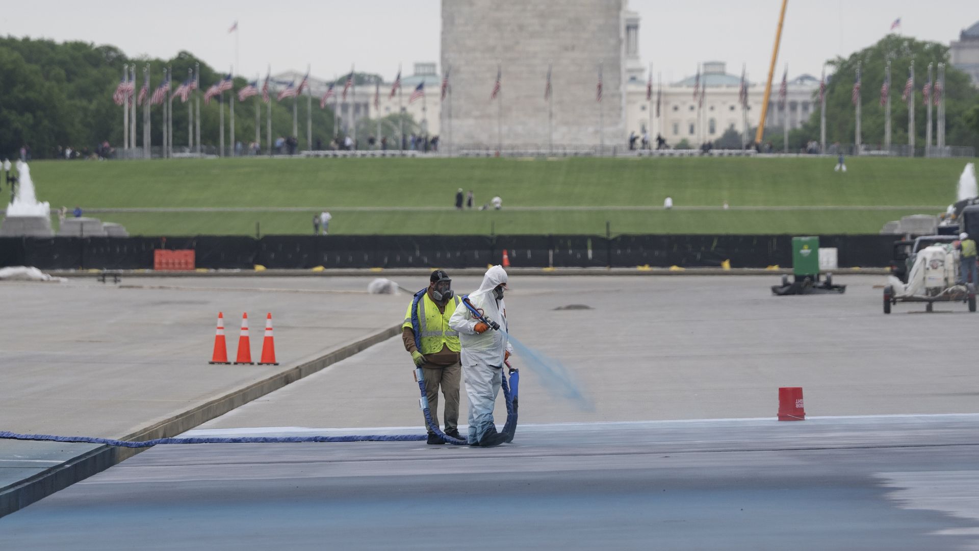 Two people in hazmat suits walk on the Reflecting Pool in the foreground, flanked by orange cones. Behind them, a green lawn, a row of American flags, and a tall obelisk Washington Monument