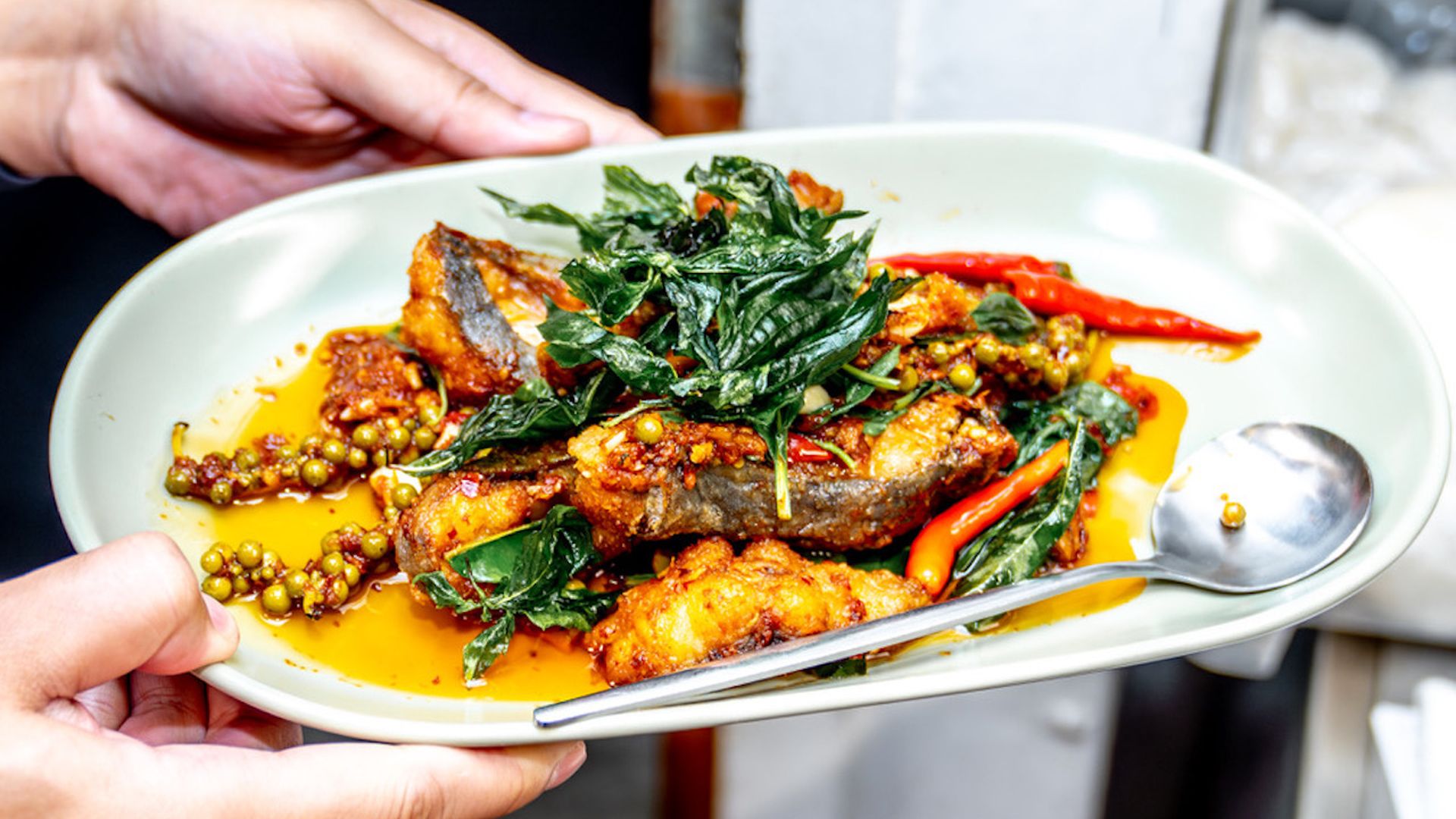 A white oval plate held by two hands, with crispy fried fish, leafy greens, red chilies, and green peppercorns in a bright orange sauce; a spoon rests on the edge.