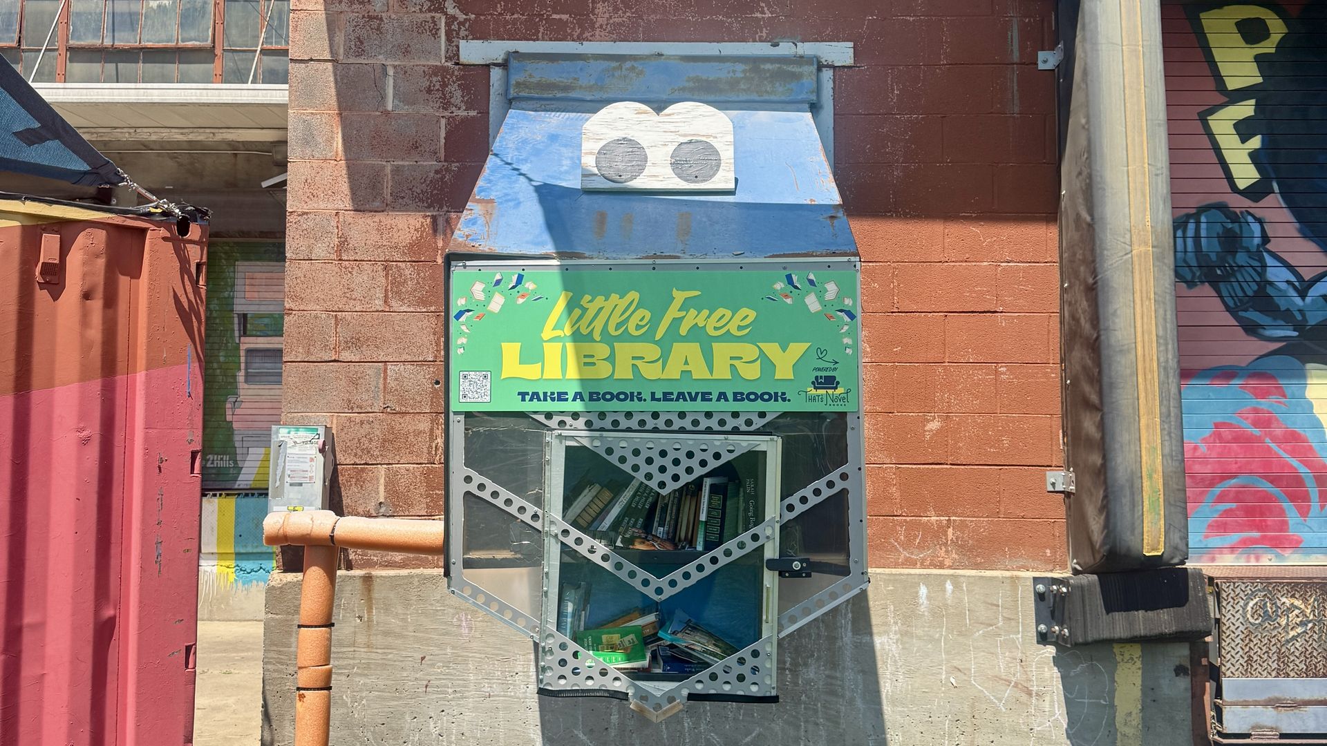 Green Little Free Library on a red brick wall under a blue metal marquee; sign reads Little Free LIBRARY and Take a book, leave a book. Graffiti and pipes frame the urban scene.