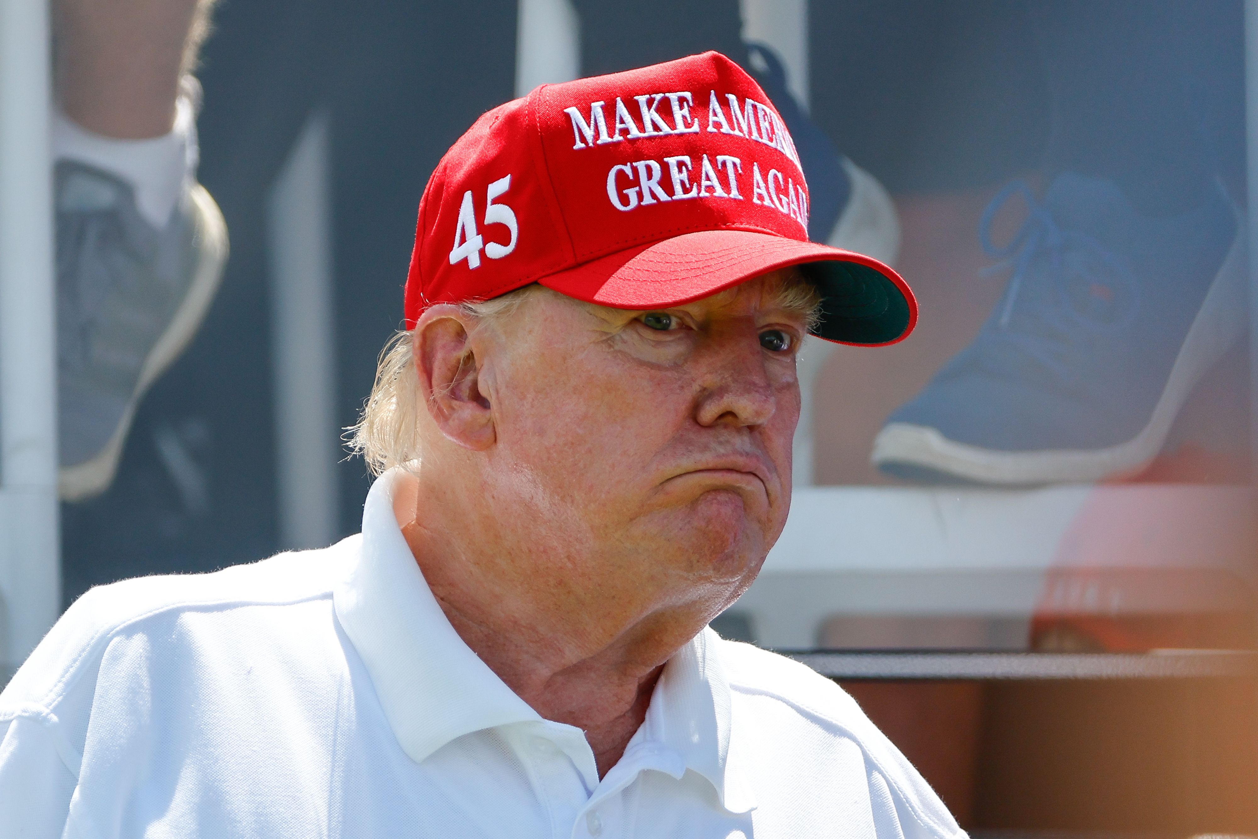 Donald Trump, wearing a white polo shirt and a red 'Make America Great Again' hat, frowns at something in the distance. 