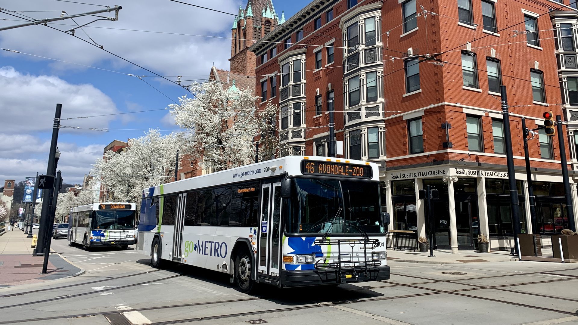 Red brick corner building with ornate trim on a city street, trolley wires overhead. A goMETRO bus, blooming white trees, and a distant church spire against a blue sky.