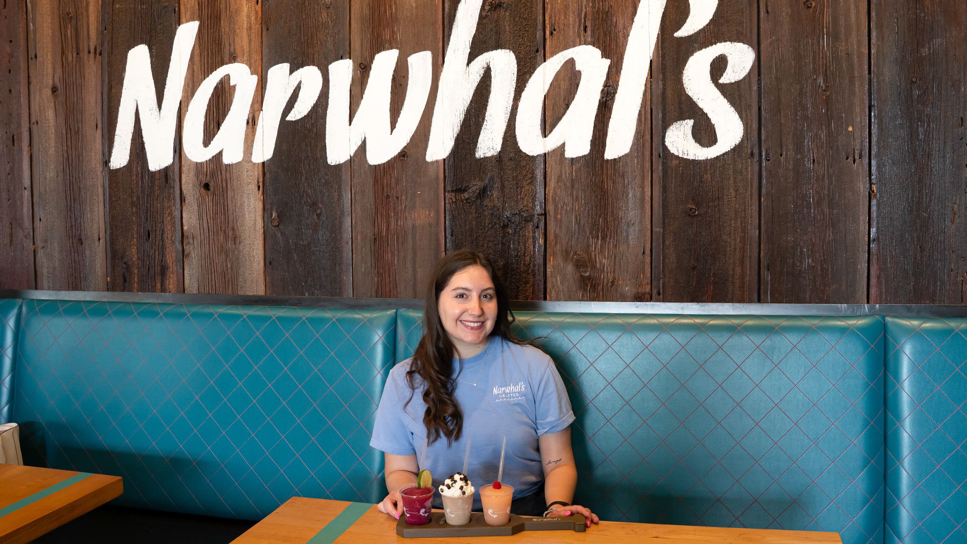 Smiling woman in a light blue shirt sits at a booth with a dark wood wall and a large white cursive "Narwhals" sign behind her; a tray of colorful milkshakes and desserts sits on the table.