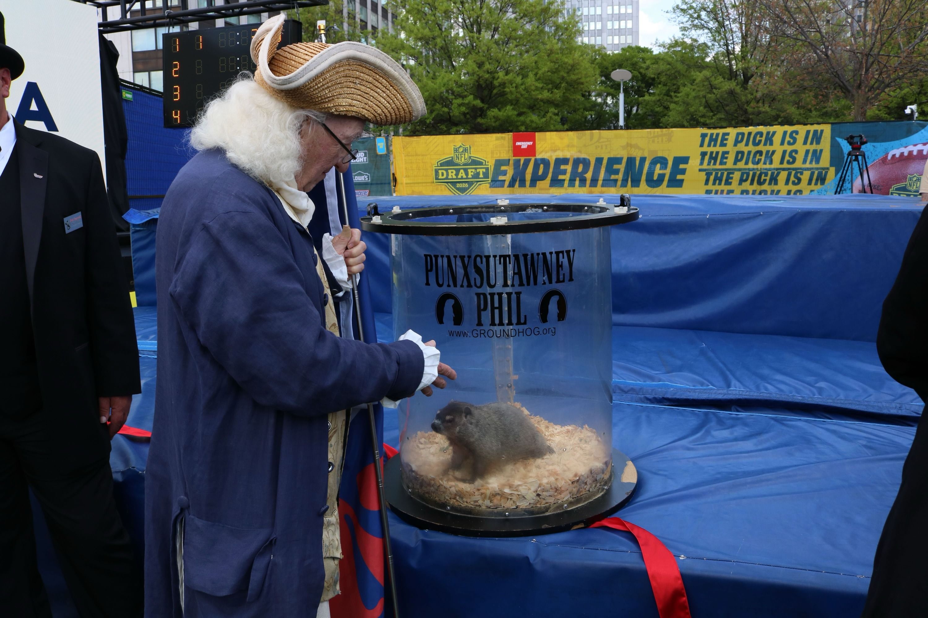 An elderly man in a blue coat, white wig, and a tricorn hat leans toward a clear cylinder housing Punxsutawney Phil during an outdoor event, with blue stage covers and a bright banner.