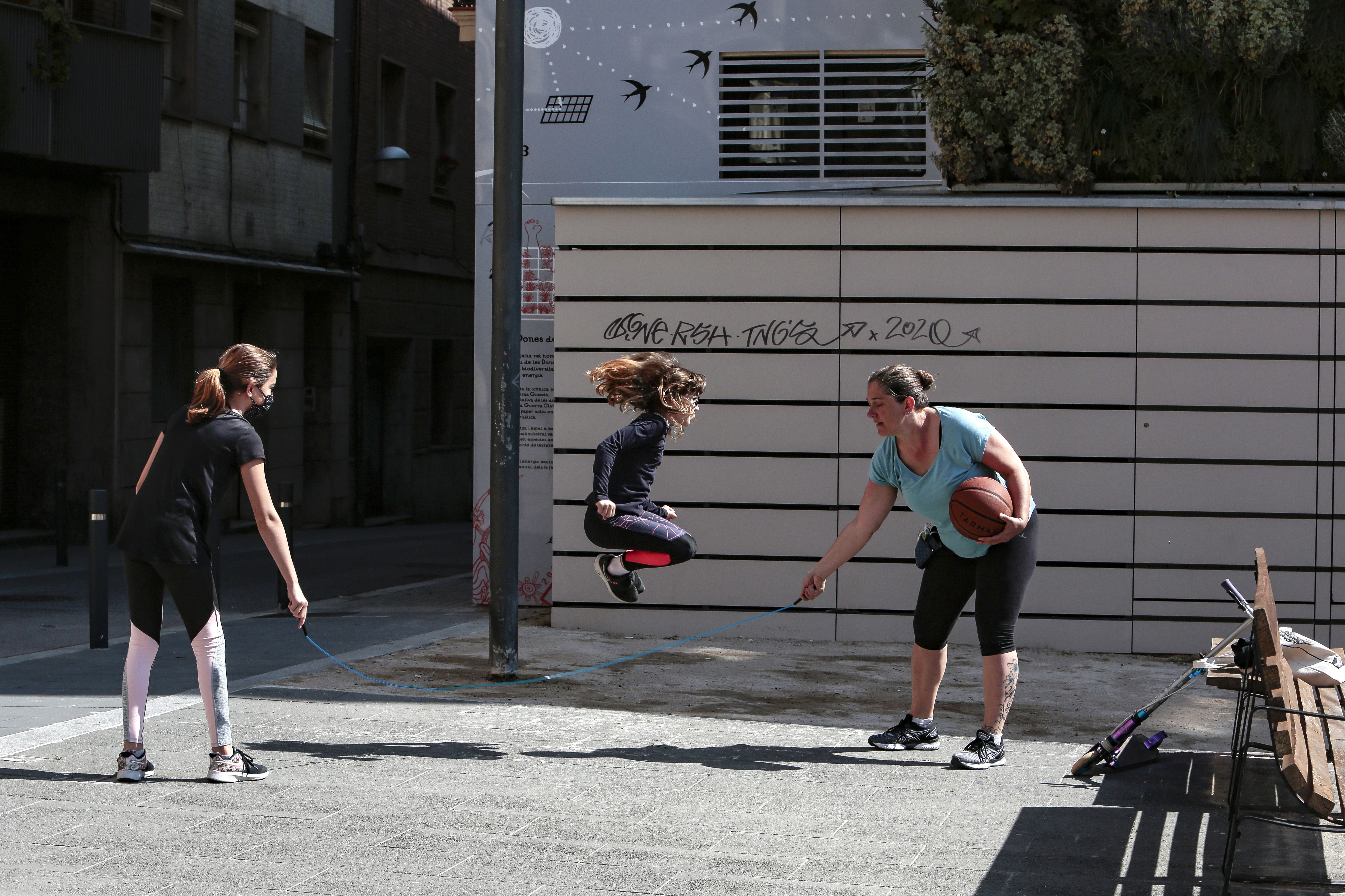 In this image, a girl jumps rope that is held by two women on a sidewalk