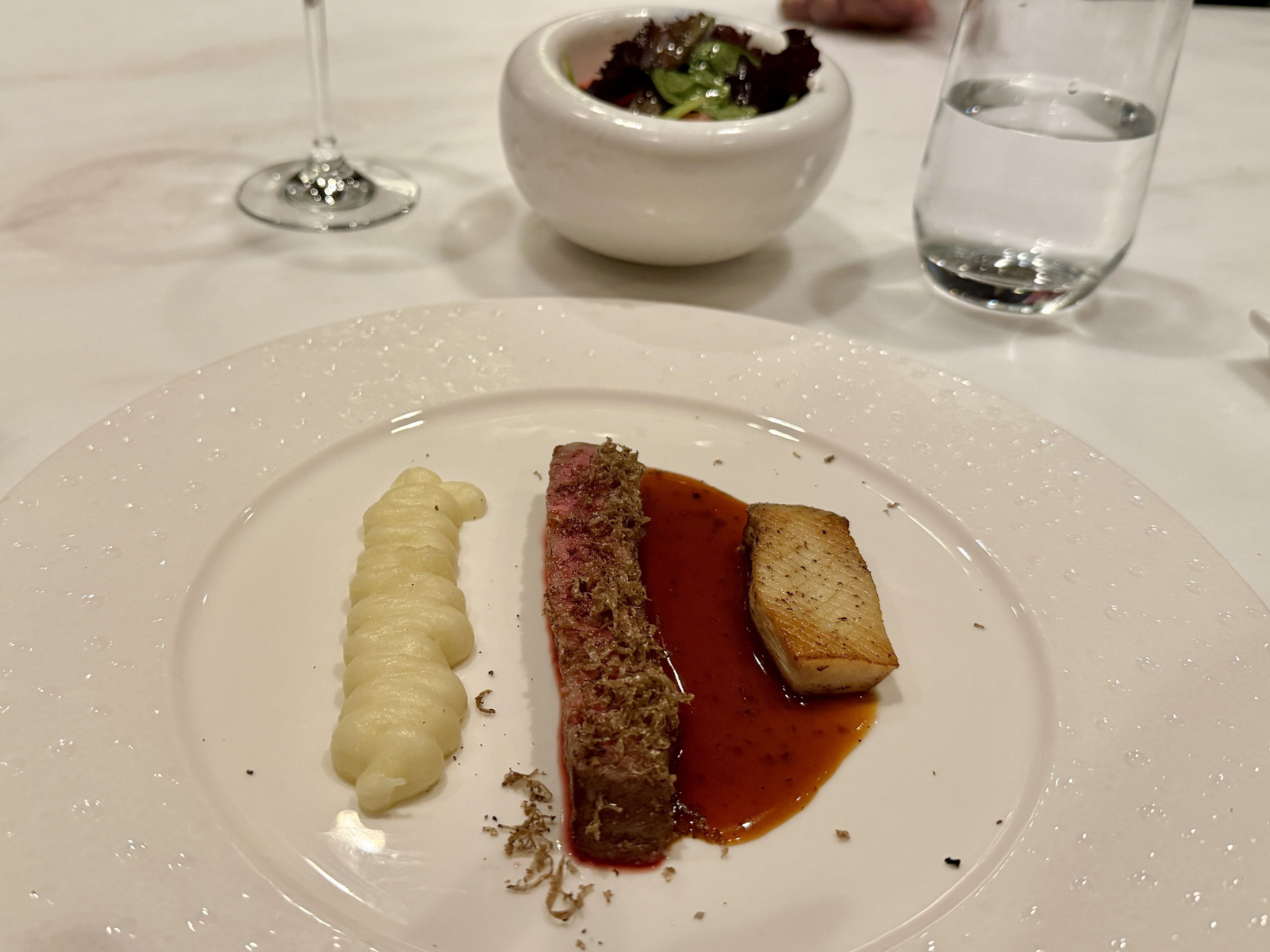 Fine dining plate with a slice of medium-rare steak topped with grated truffle, a dollop of creamy white mashed potatoes, and a rectangular piece of seared food with reddish-brown sauce, set on a white table.