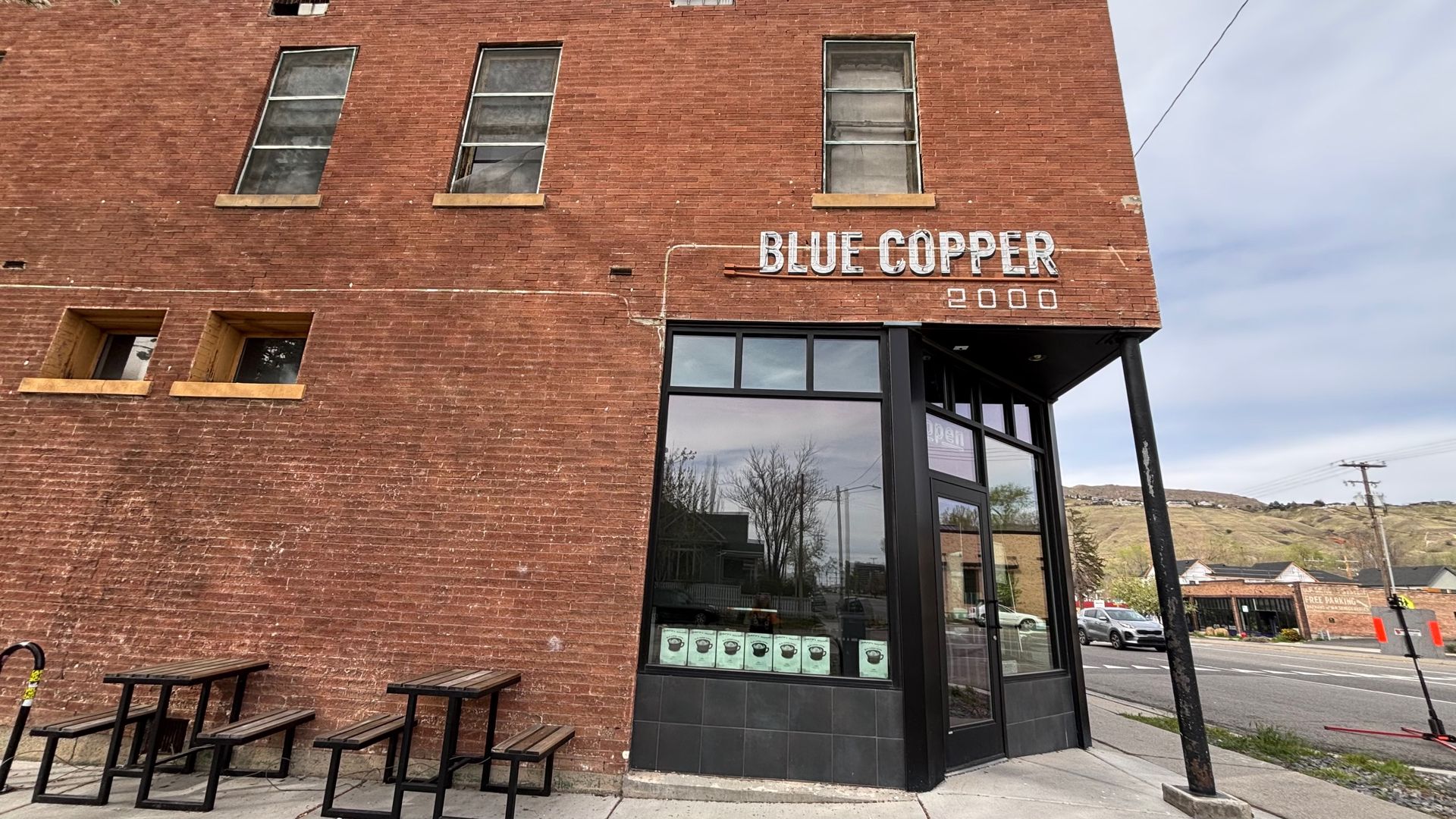 Red brick building with a white "BLUE COPPER" sign and the number 2000. Large storefront window; outdoor wooden picnic tables on the sidewalk. Street, distant hills, and blue sky.