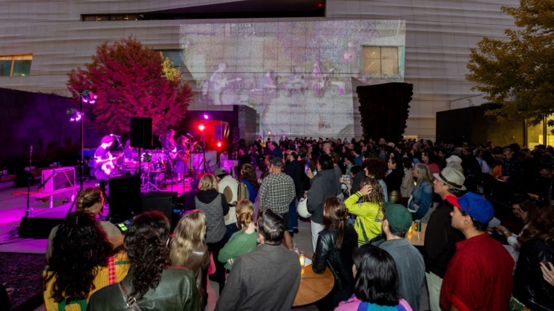 Outdoor concert at dusk with a purple-lit band on a stage to the left, a large crowd facing them, modern building wall projection in the background, and trees on either side.