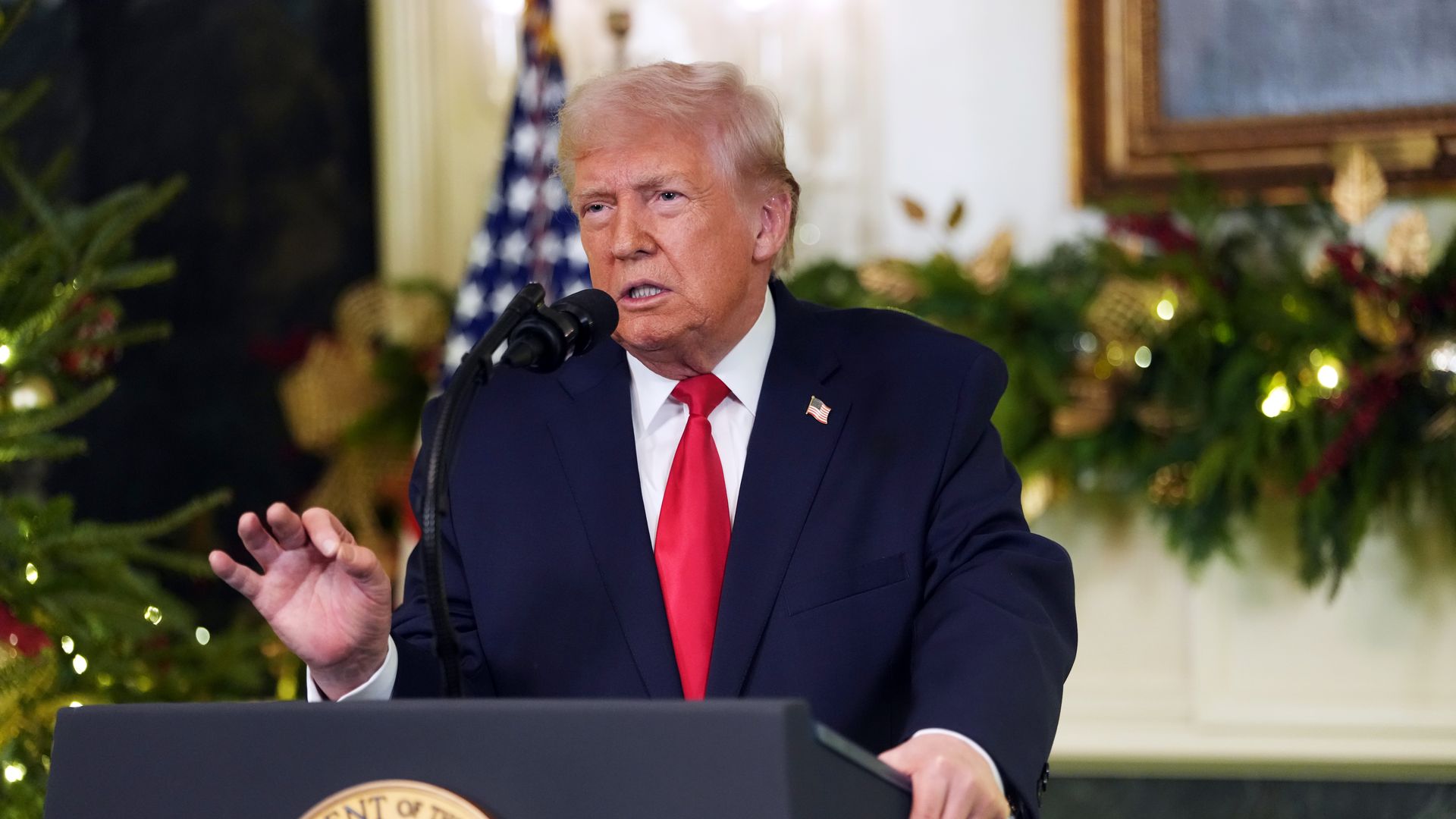 President Trump — wearing a navy suit, a red tie, a white collared shirt and an American flag pin — holds his hand up as he speaks into a microphone on a podium in a room in the White House decorated for the holidays. 