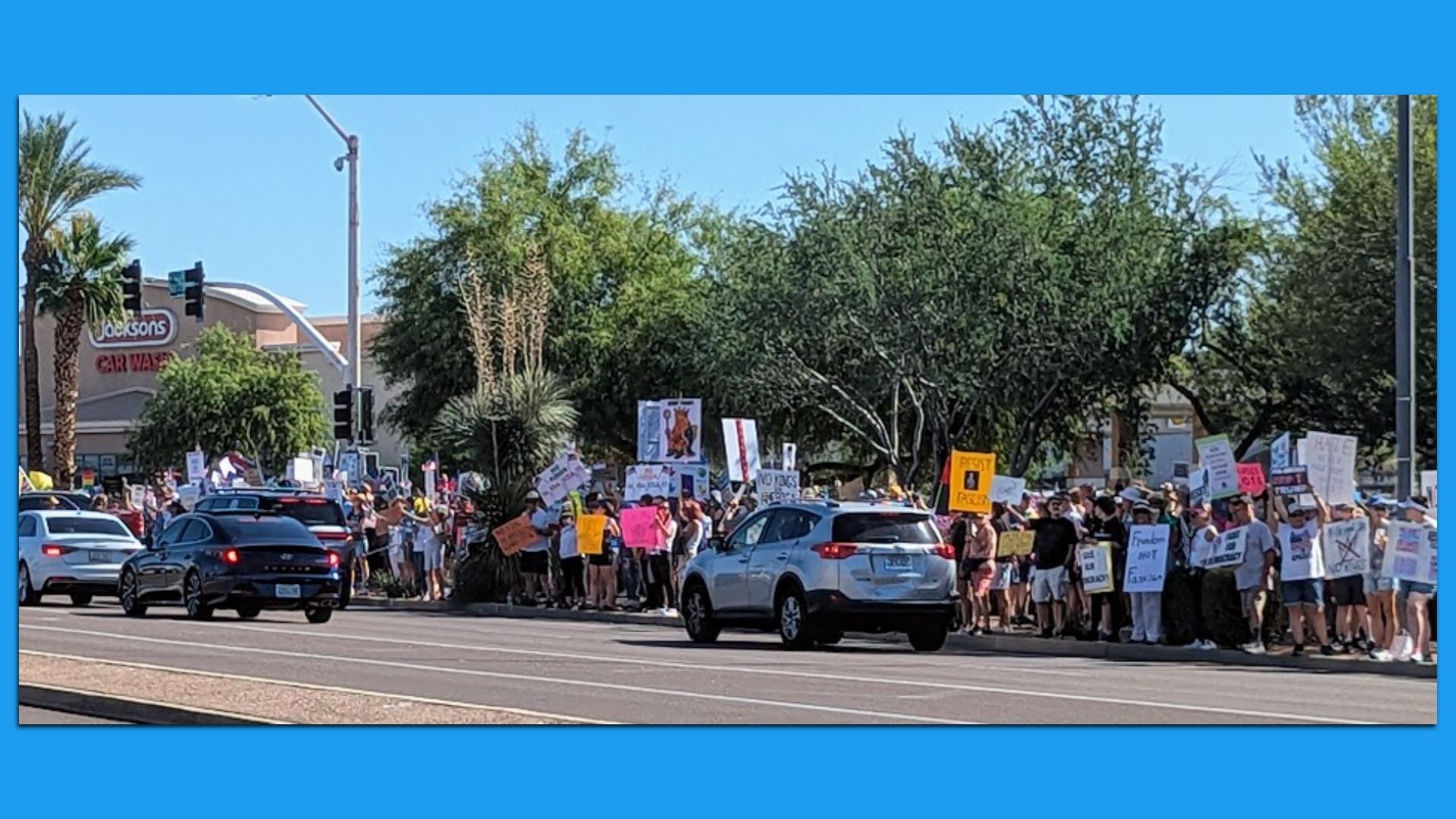 Protesters waving signs line a sidewalk as cars drive past. 