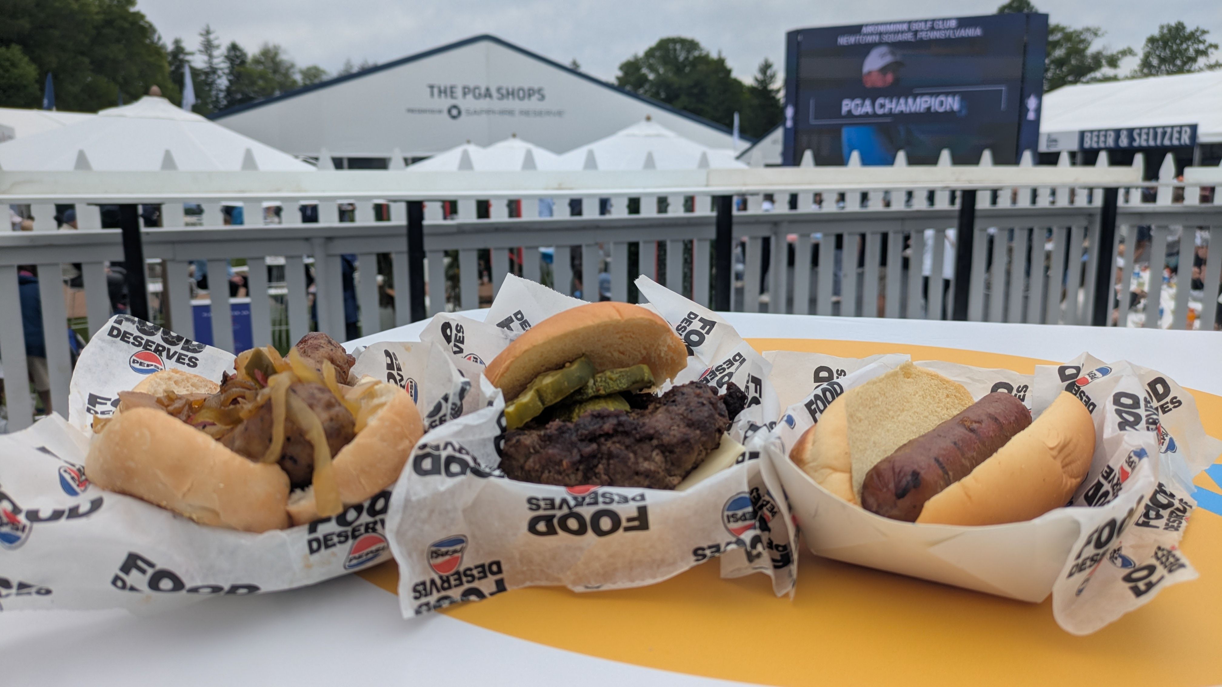 Close-up of hot dogs and grilled snacks on a table, wrapped in branded paper. In the background, white railing and tents, with a large scoreboard reading "PGA CHAMPION" at a golf event.