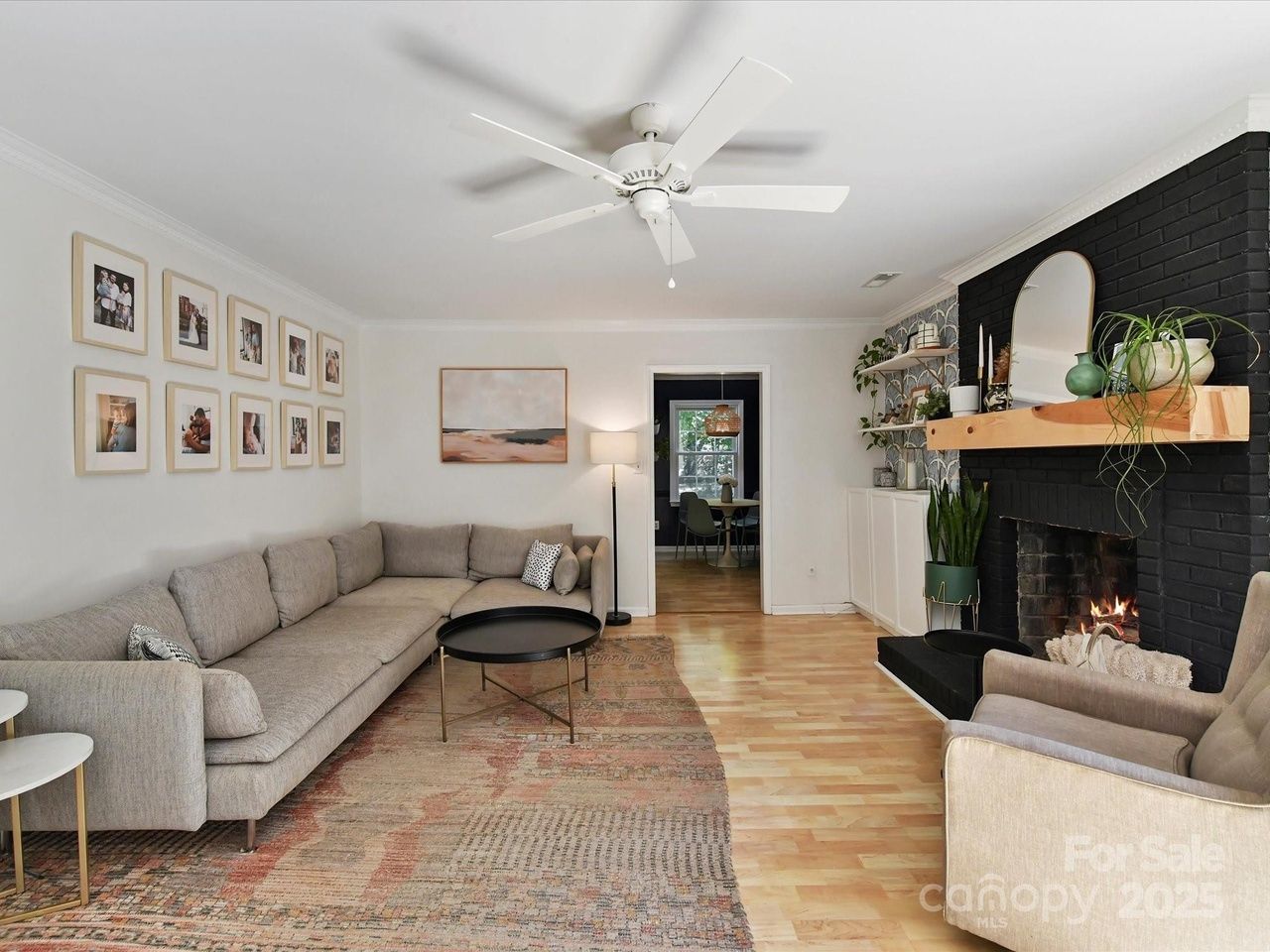 Living room with a gray sectional sofa, black round coffee table, beige armchair, colorful rug, black brick fireplace with plants and mirror, and white ceiling fan.