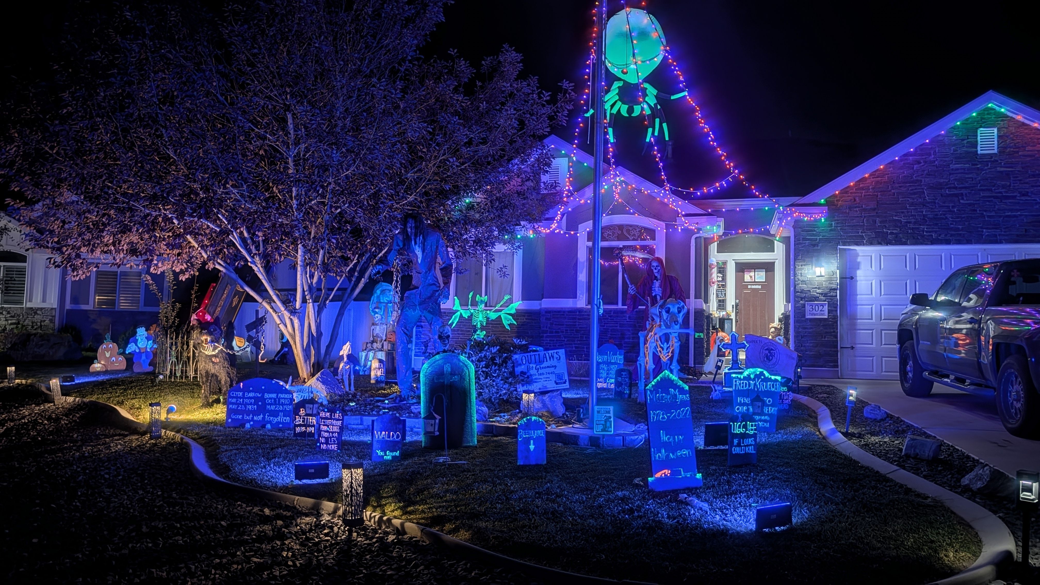 Nighttime suburban Halloween yard scene: house outlined with multicolored string lights, eerie tombstones, skeletons and props, pumpkins, trees, and a truck SUV in the driveway.