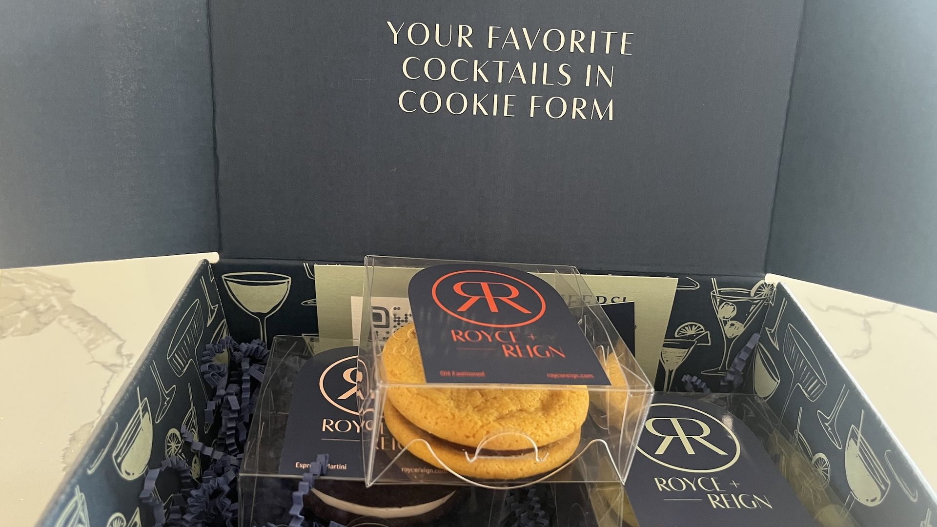 open purple box with stack of 3 cookies in plastic boxes in it. The box reads: Your favorite cocktails in a box.