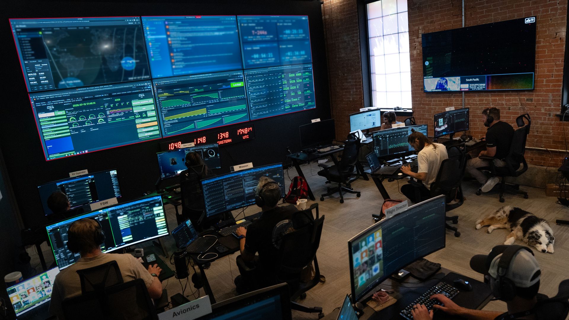 Control room with a wall of large monitors displaying dashboards, maps, and graphs. Operators at desks with laptops and headsets in a brick-walled space, with dogs resting on the floor.