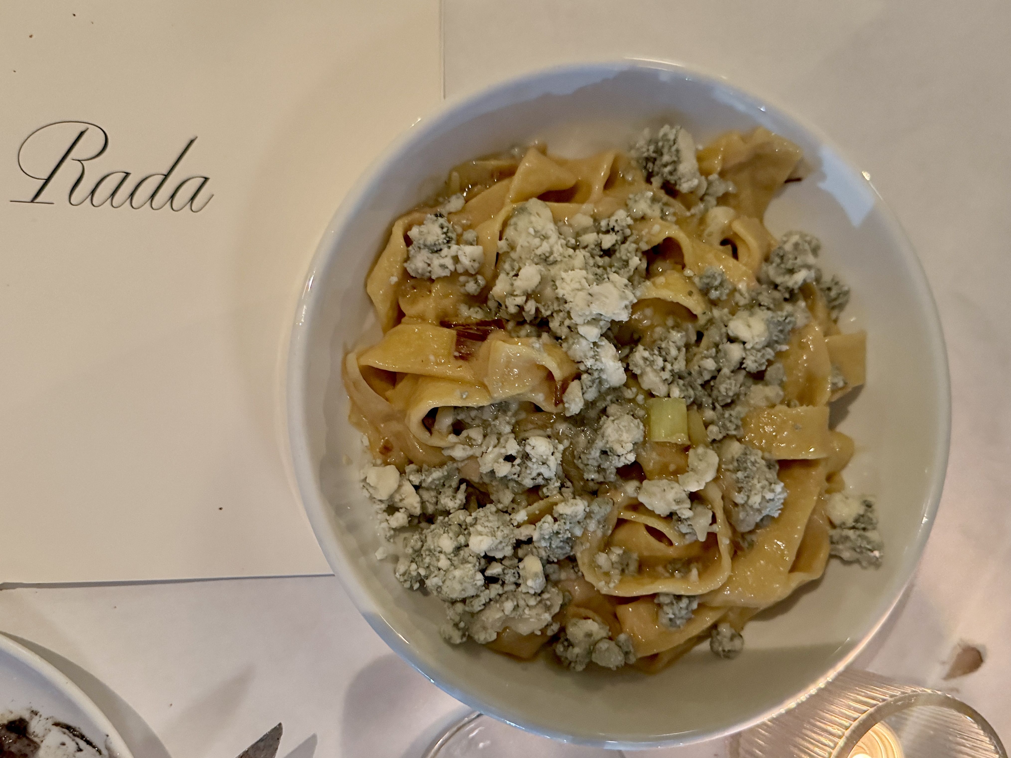 White bowl of wide pasta with crumbled blue cheese on top, placed on a white table next to a paper with the word "Rada" in script.